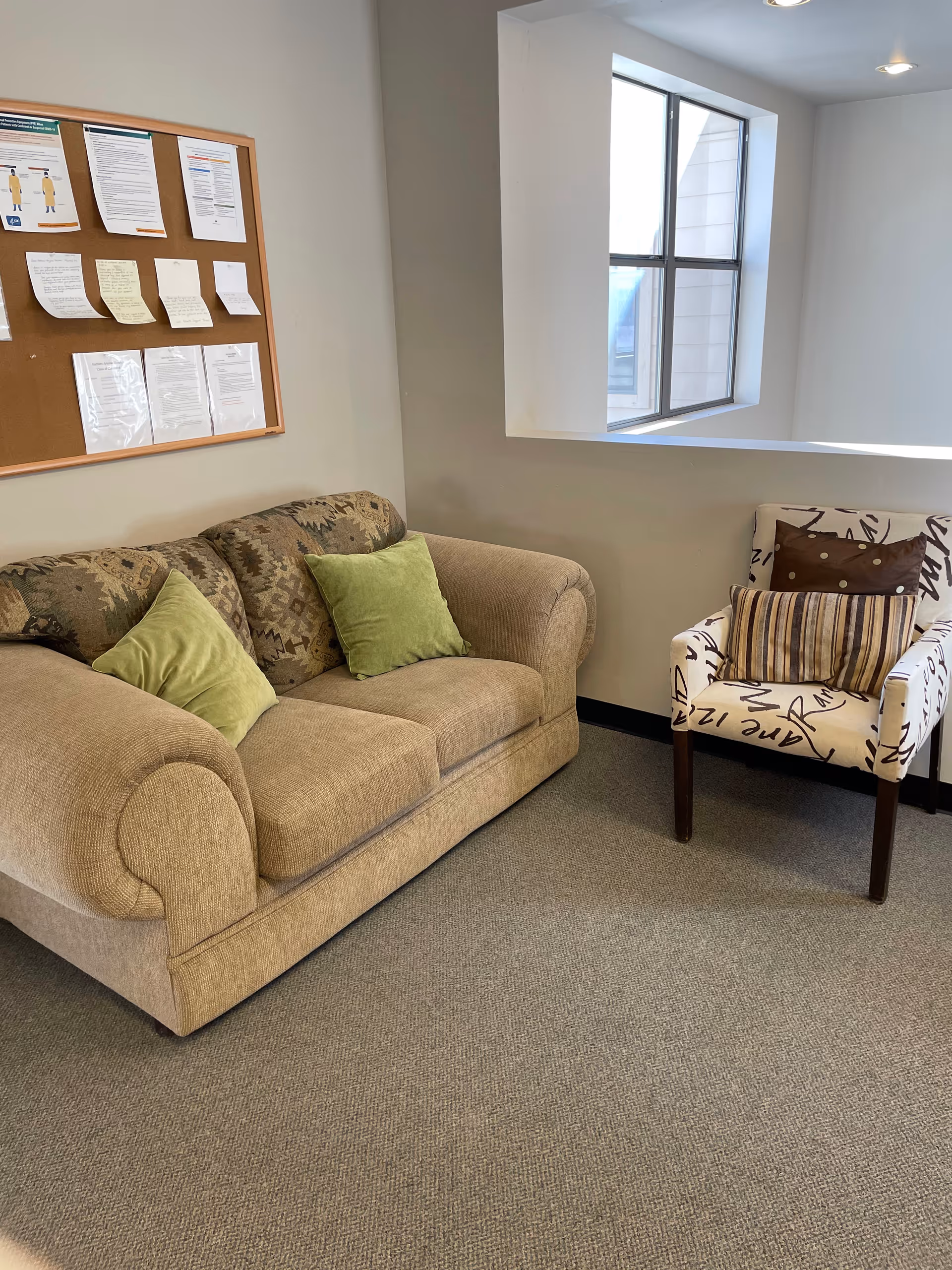 Cozy living area with beige couch, patterned armchair, cushions, and bulletin board.