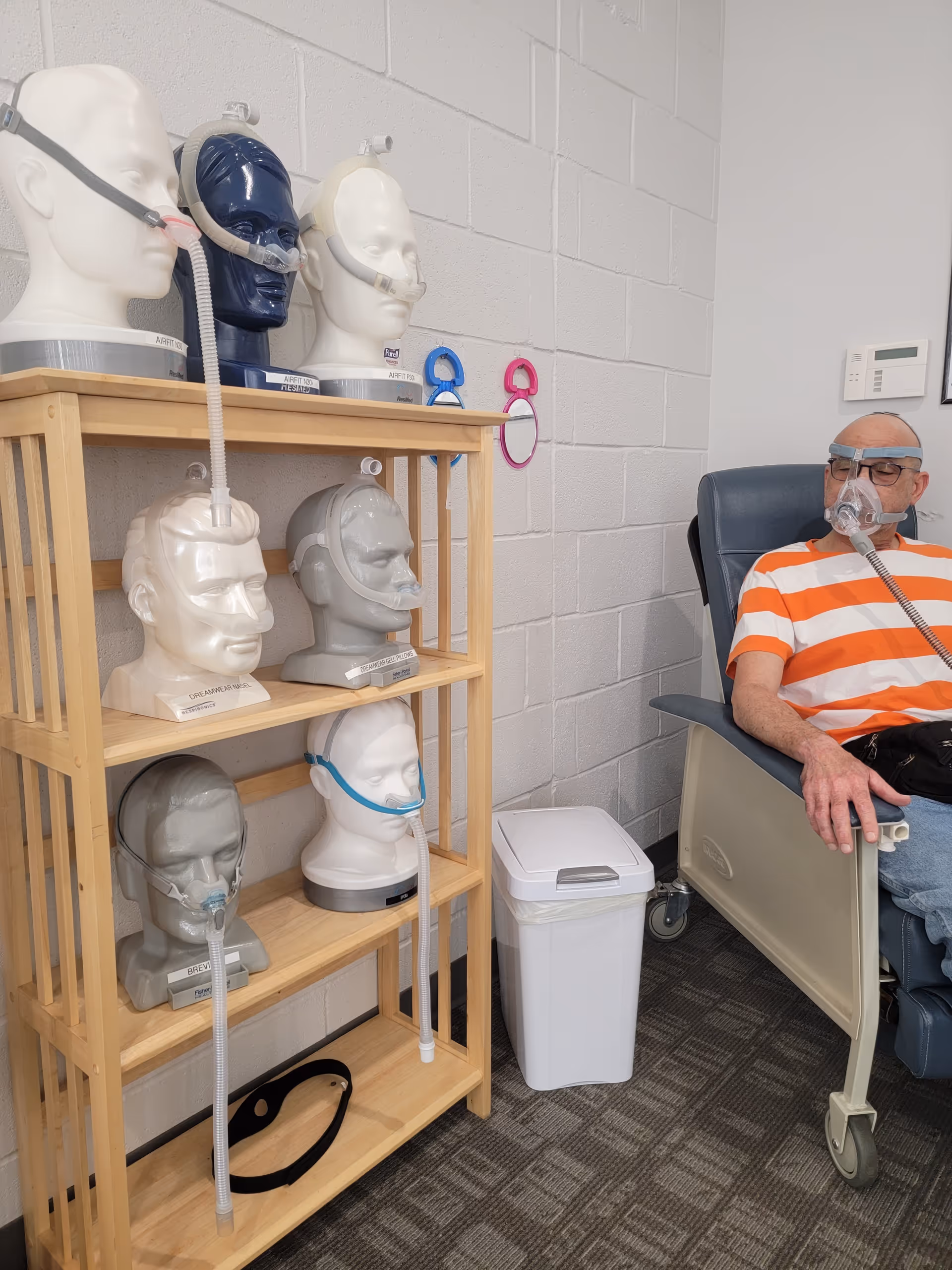 Senior man wearing a CPAP mask sits beside shelves of oxygen masks.