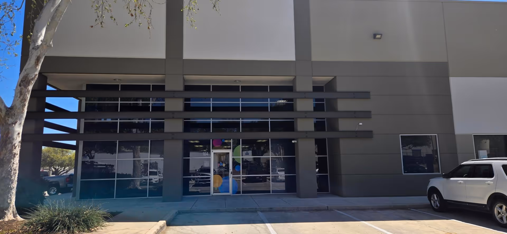 Modern facility front with glass entrance, gray exterior, and a white SUV parked outside.