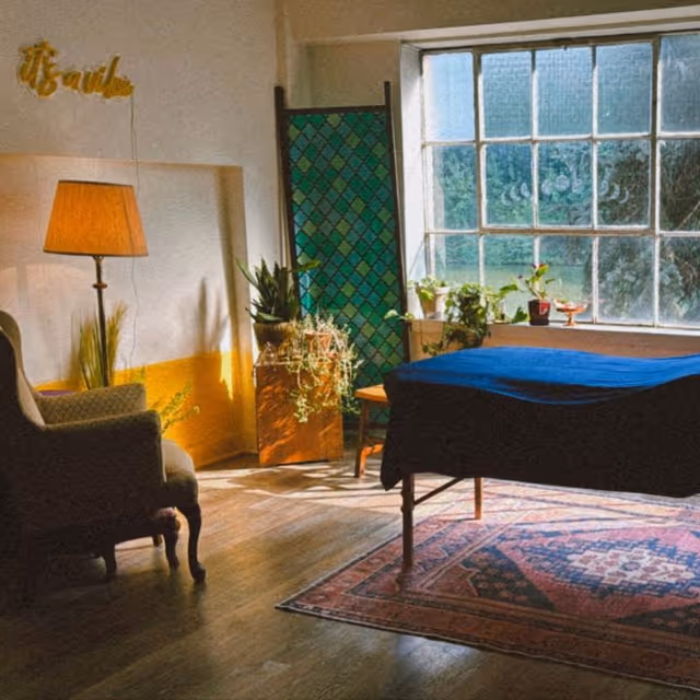 A cozy therapy room with a blue-covered table, plants, lamp, chairs, and a large window.