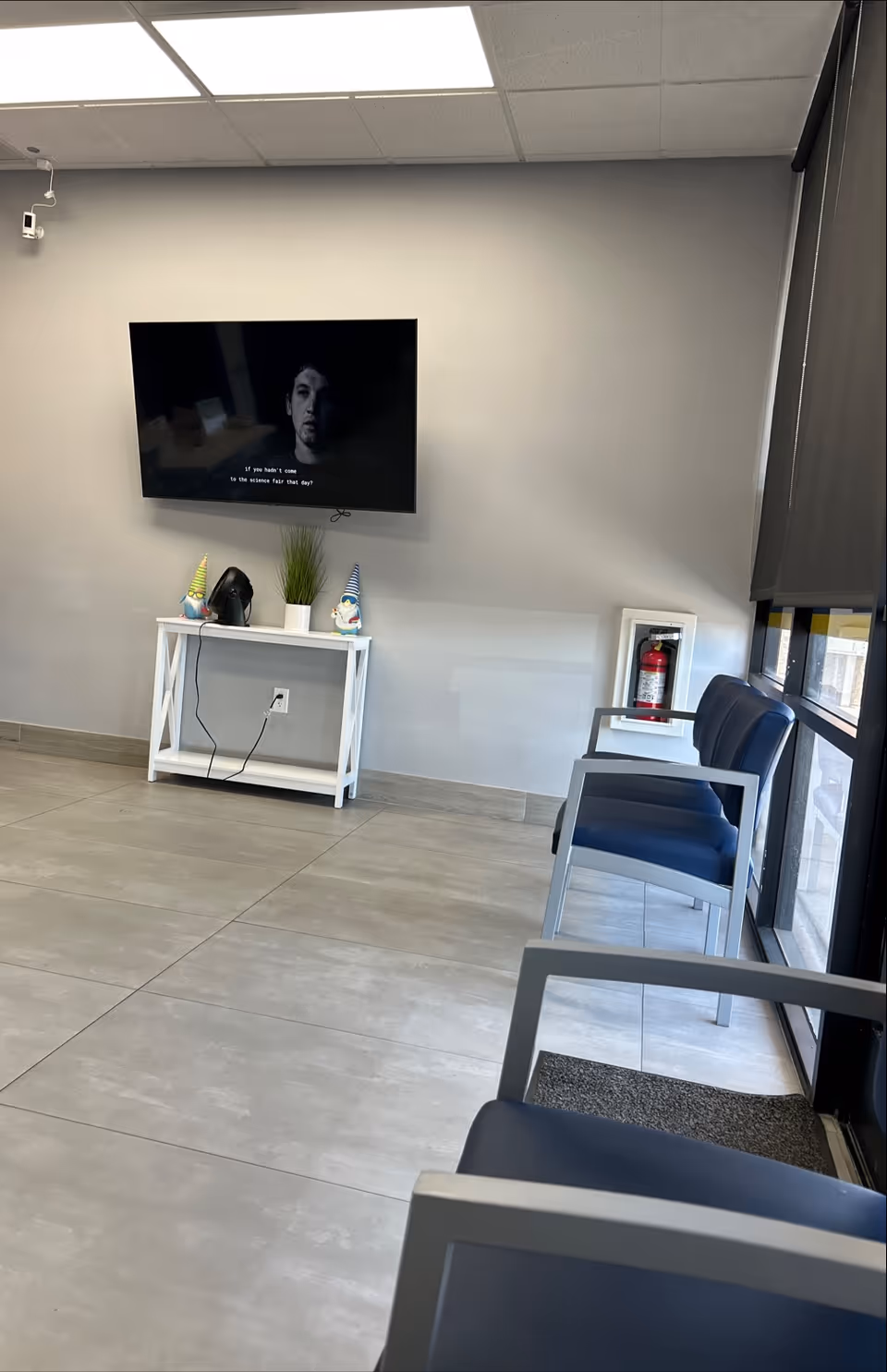 Interior waiting lounge with wall TV, white console, plants, and blue chairs