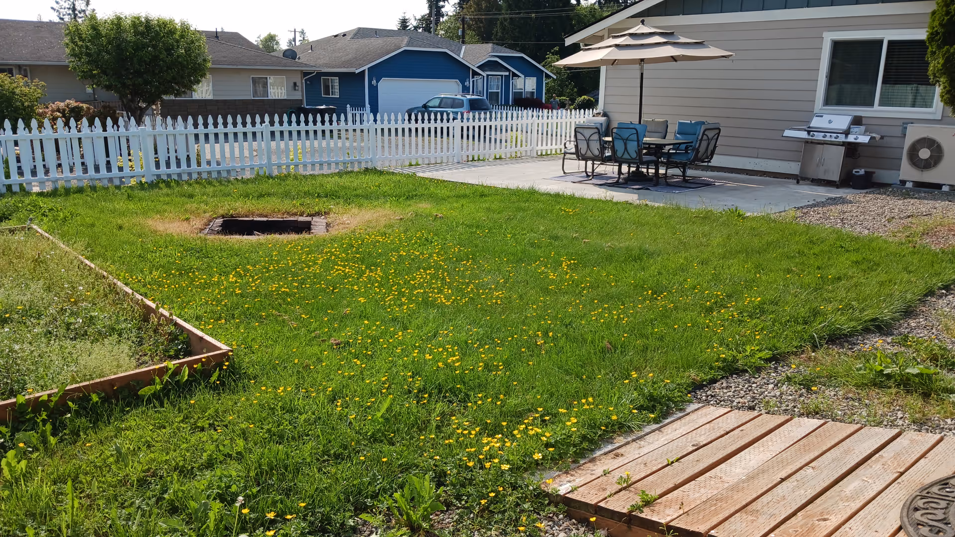 Outdoor yard with white picket fence, patio table, umbrella, and grill beside a house.