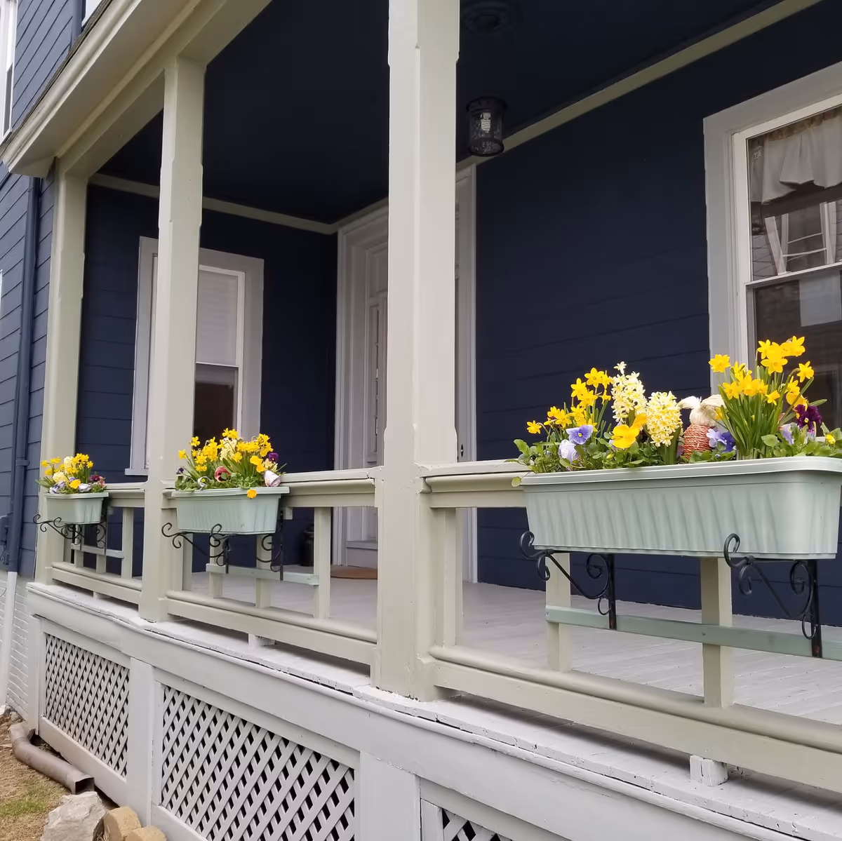 Blue house exterior with light-green porch columns and flower-filled planters on the railing.
