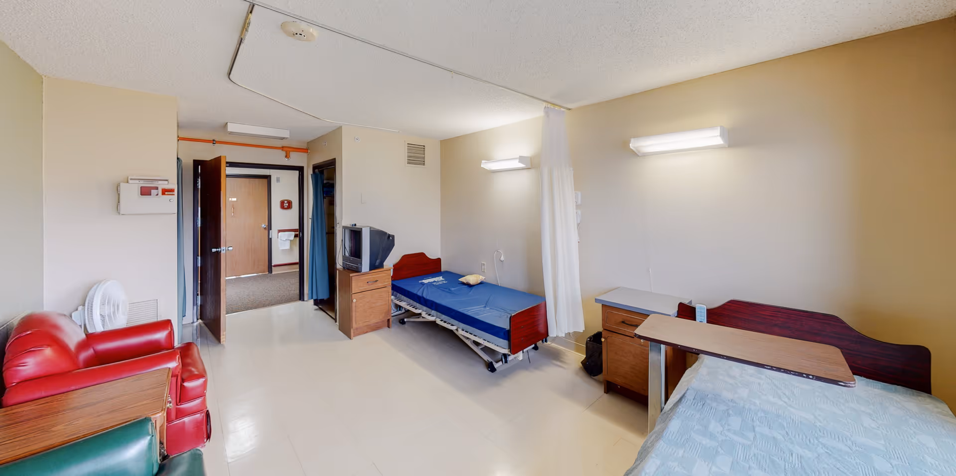 Hospital-style patient room with two beds, a red sofa, desks, and bright wall lights.