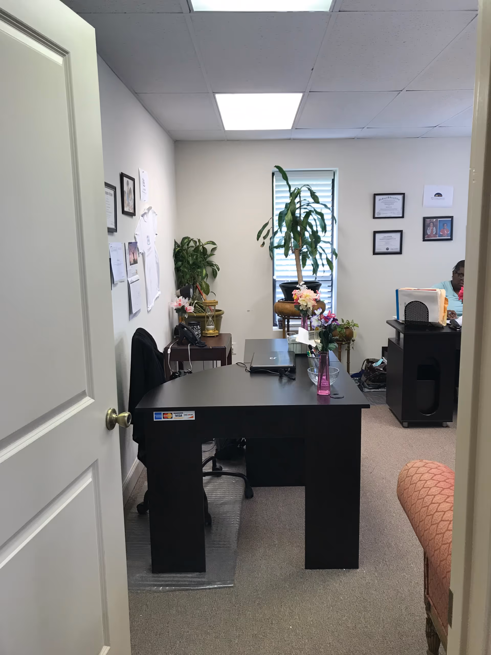 Office workspace with a large black desk, plants, frames, and a partial person.