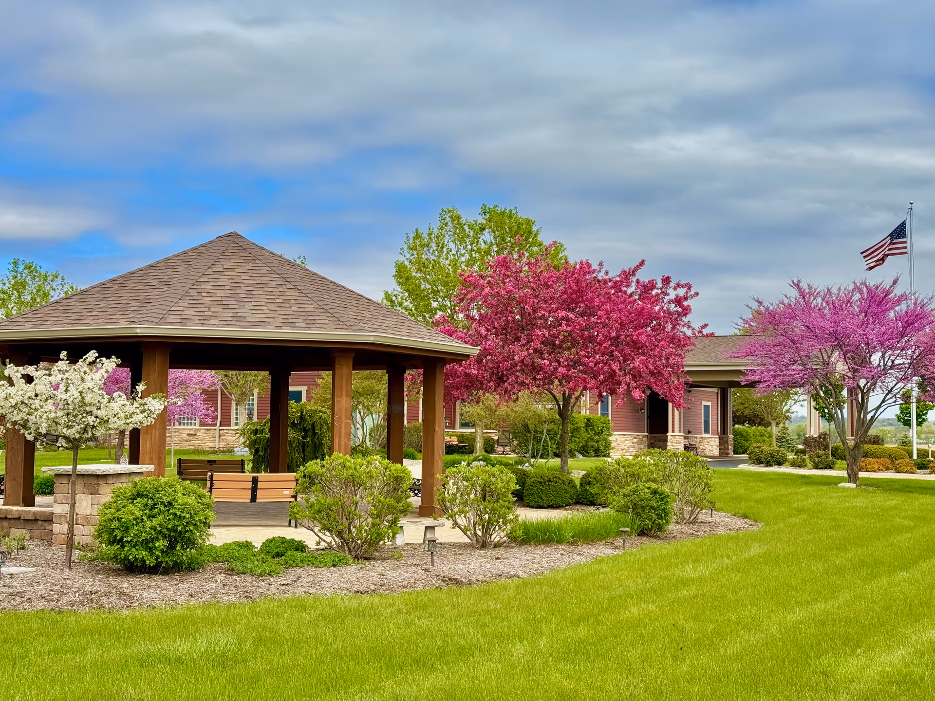 Well-kept lawn with a wooden gazebo, blooming trees, and a building in the background.