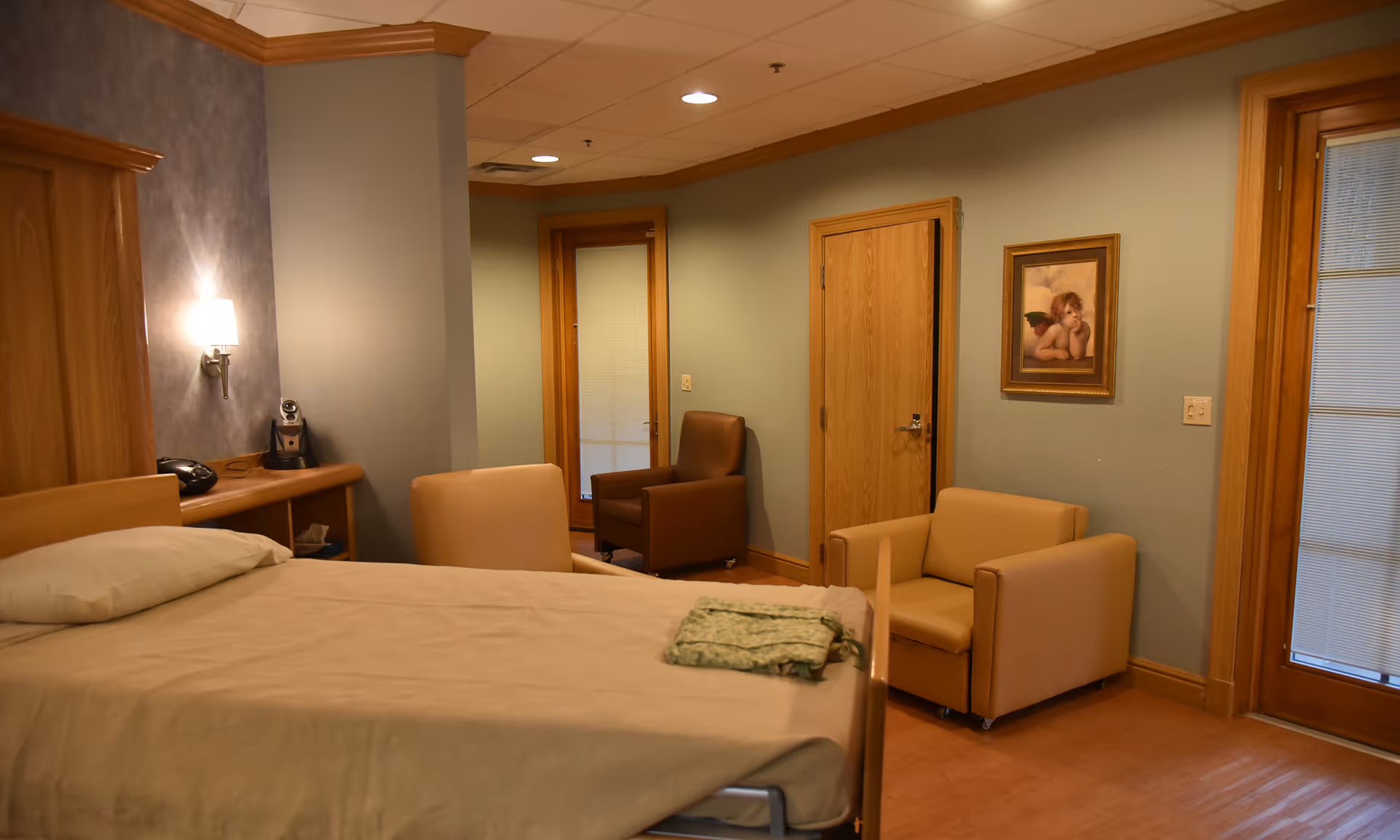 A hospice bedroom with a bed, multiple chairs, doors, and framed art.