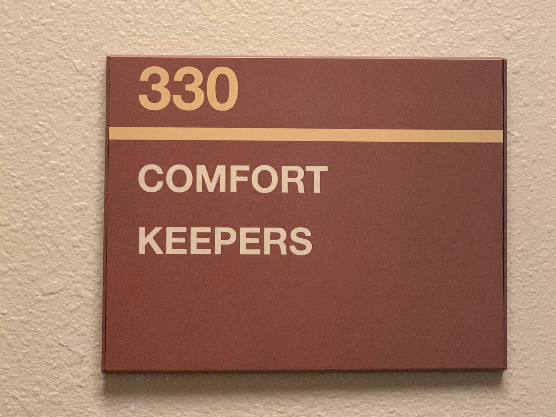 Maroon hallway sign reading 330 Comfort Keepers mounted on textured beige wall.