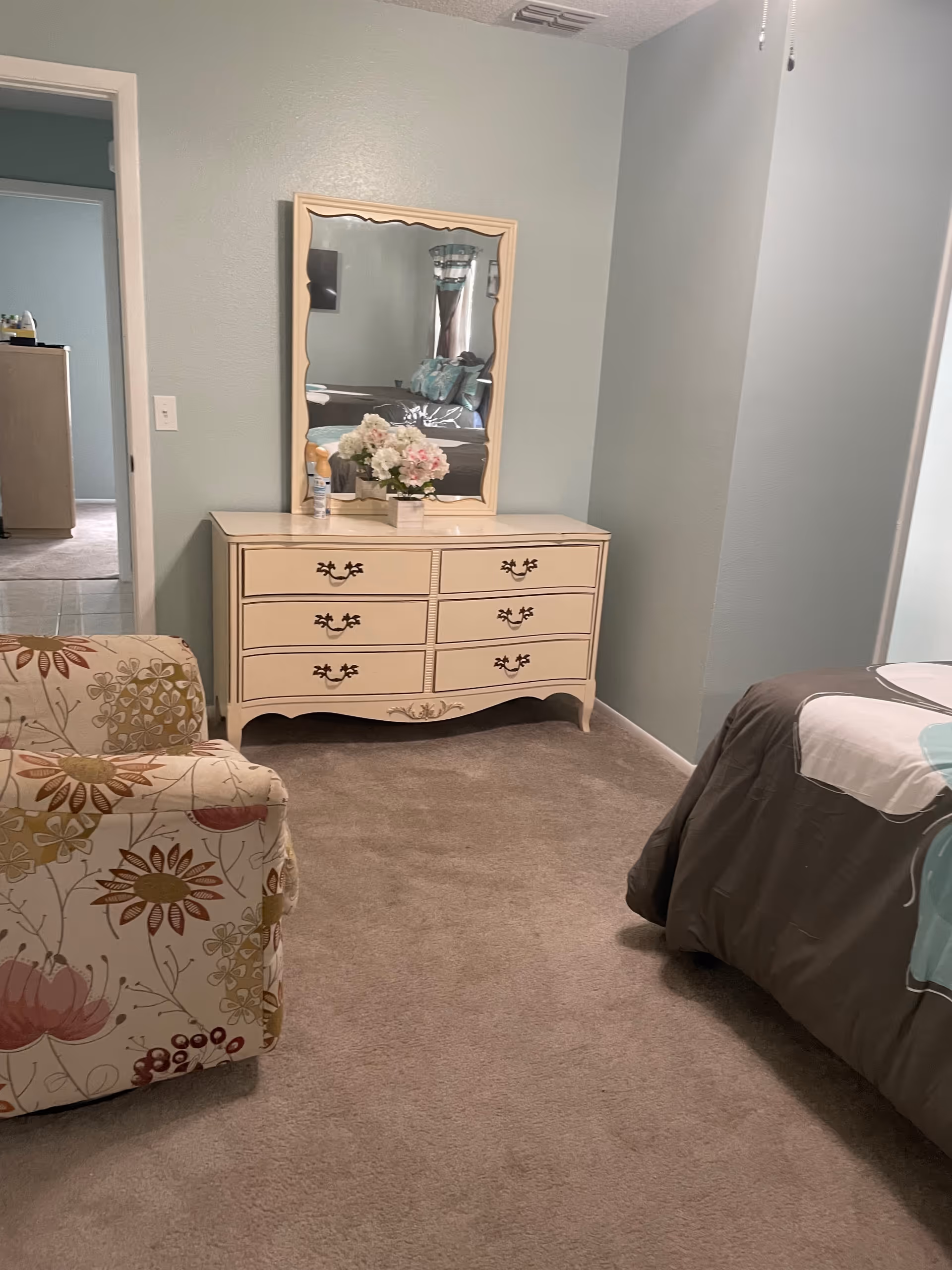Cream dresser with mirror, floral chair, and a bed in a pastel teal bedroom.