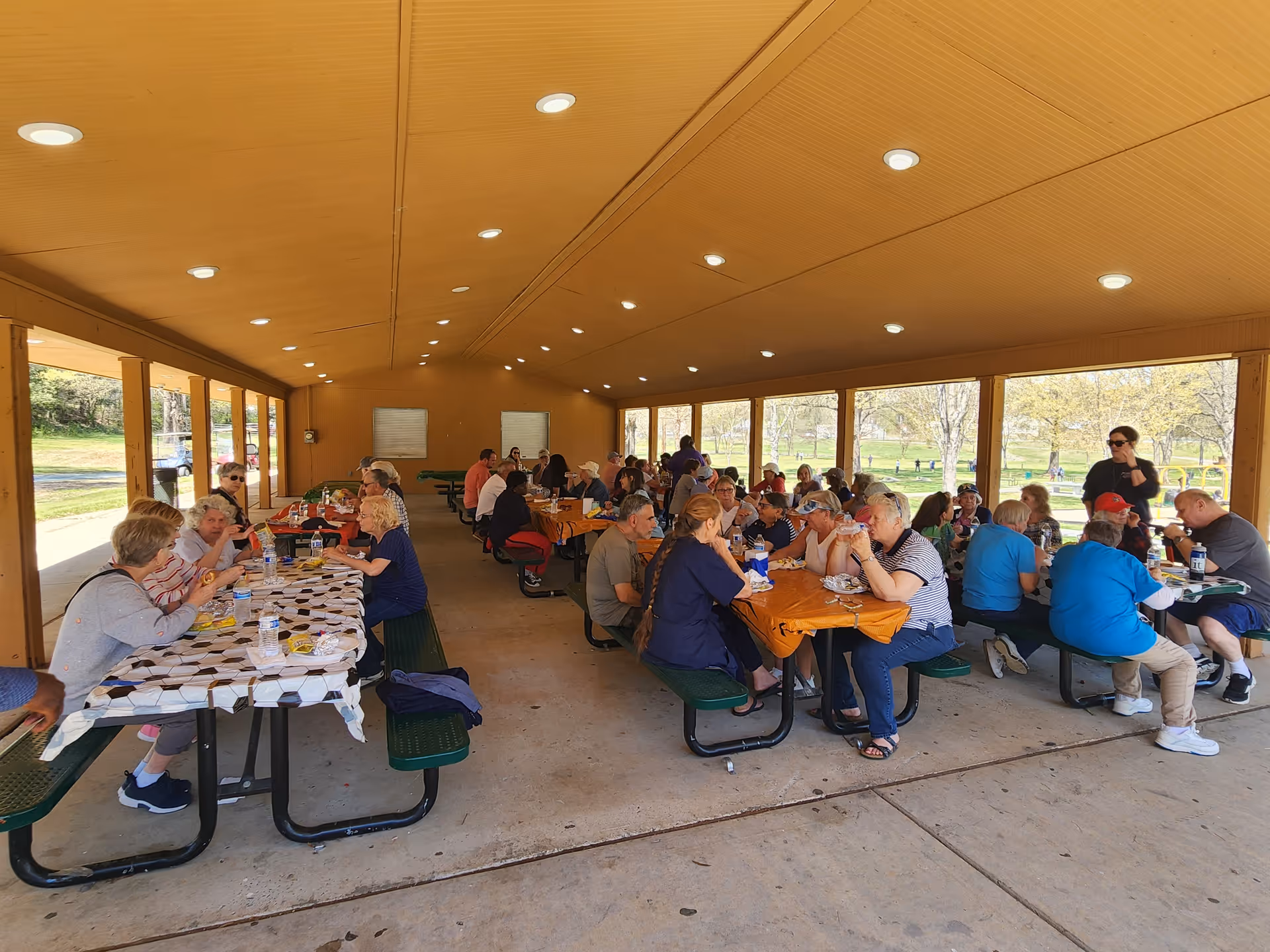 Large group of seniors dining at long picnic tables under a covered outdoor pavilion.
