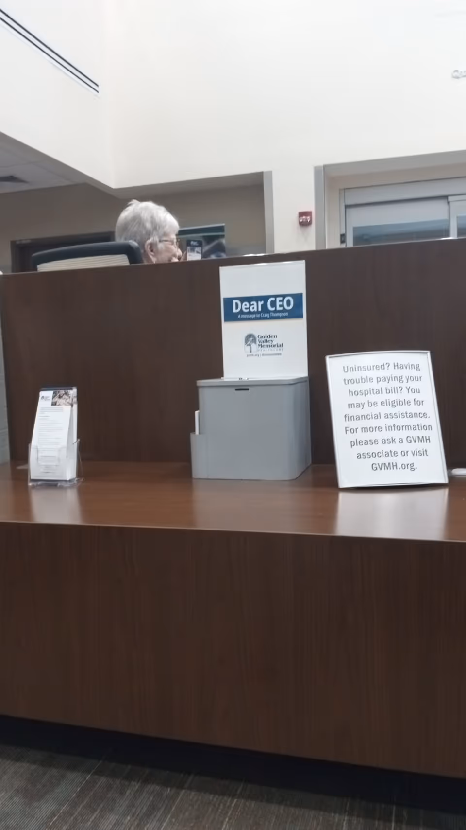 Elderly person seated behind a dark wood reception desk in a healthcare facility lobby.