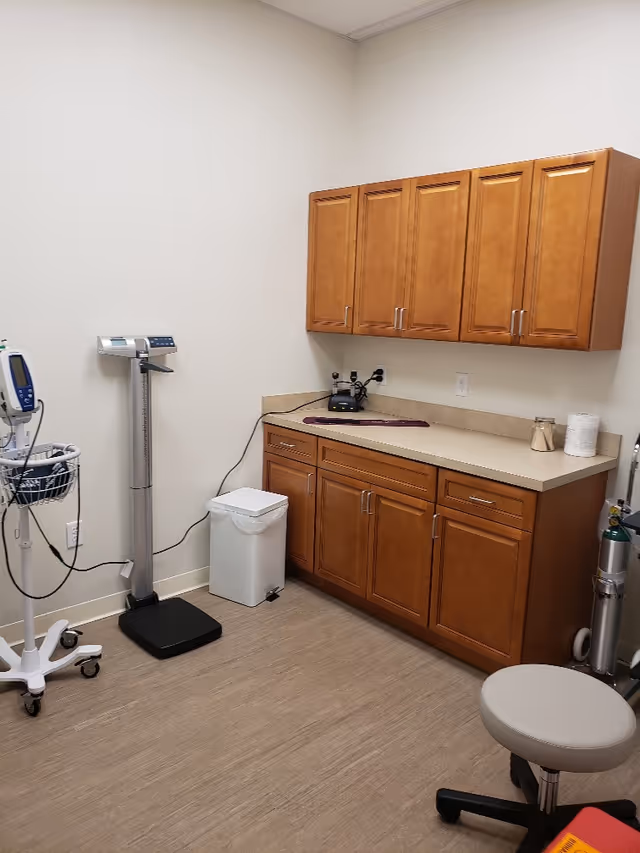 Medical examination room with wooden cabinets, counter, scale, and clinic equipment.