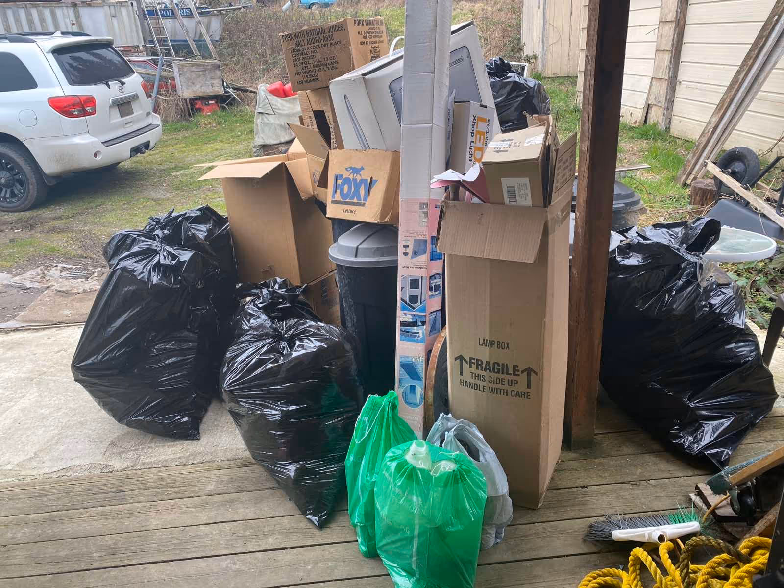 Outdoor clutter with overflowing cardboard boxes and black trash bags near a parked car.