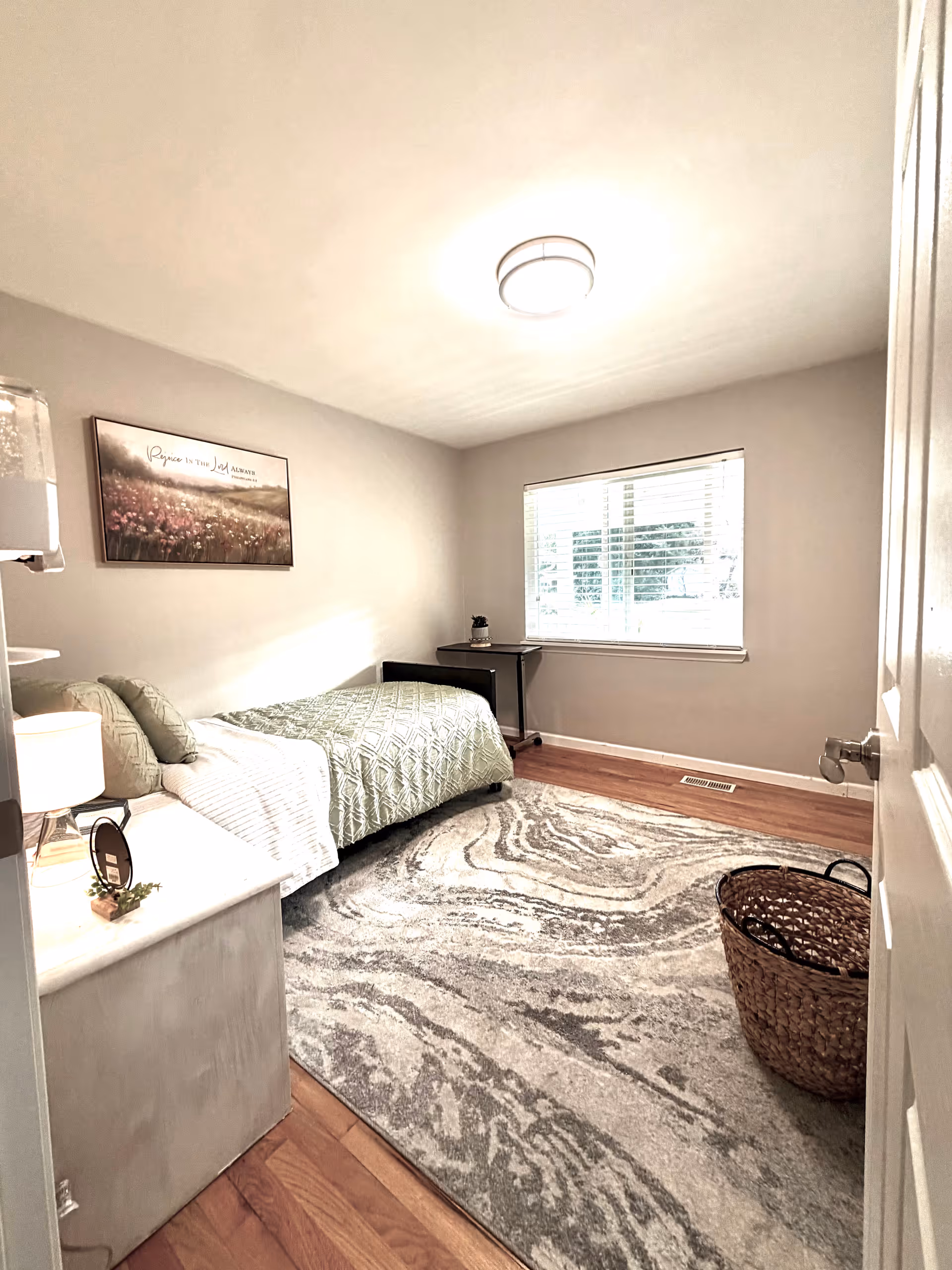 A furnished bedroom with a bed, rug, nightstand, window, and woven basket.