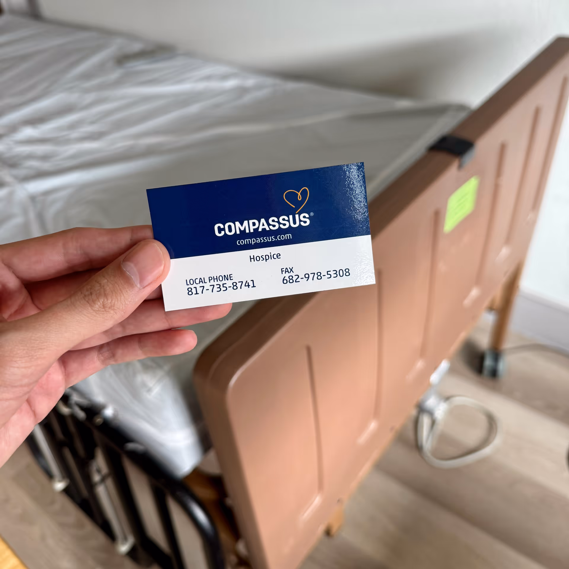 A hand holds a Compassus Hospice business card beside an adjustable hospital bed in a room.