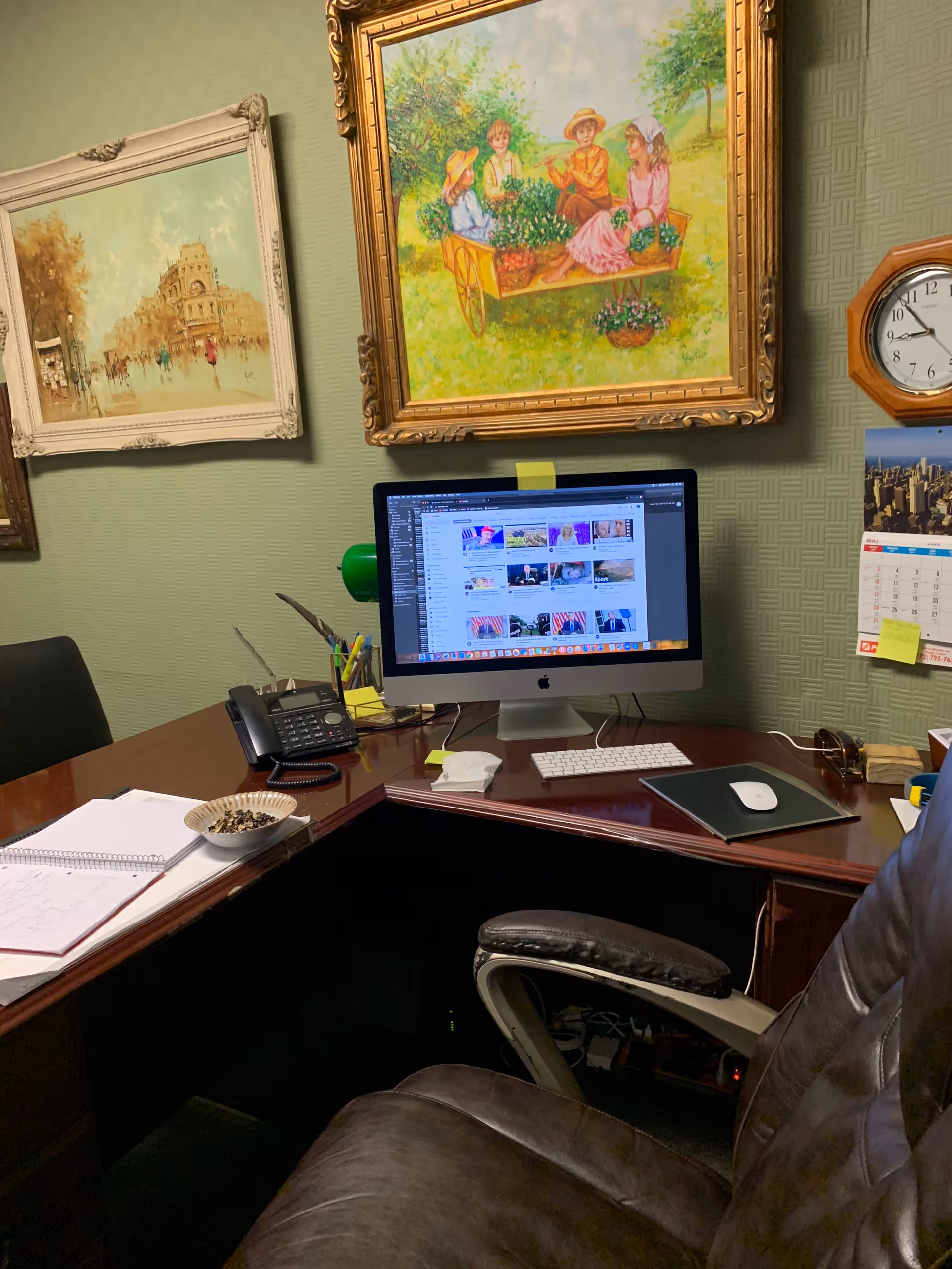 Office desk with iMac computer, office supplies, and framed paintings on a green-walled wall.