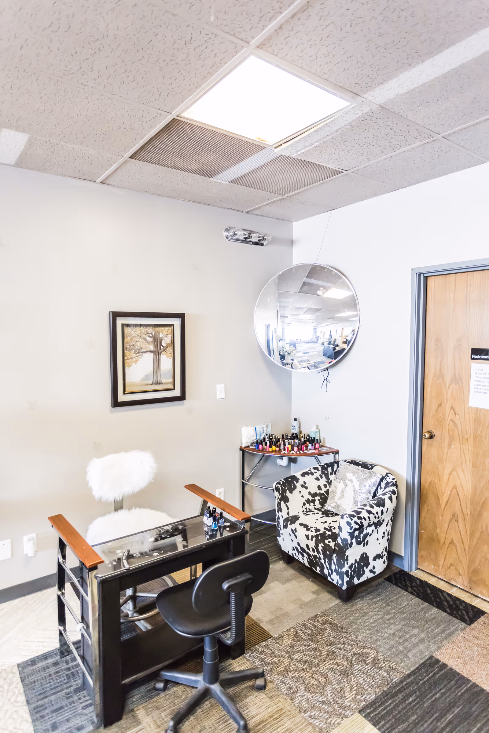 A compact salon corner with a manicure desk, round mirror, nail polish cart, and patterned seating.