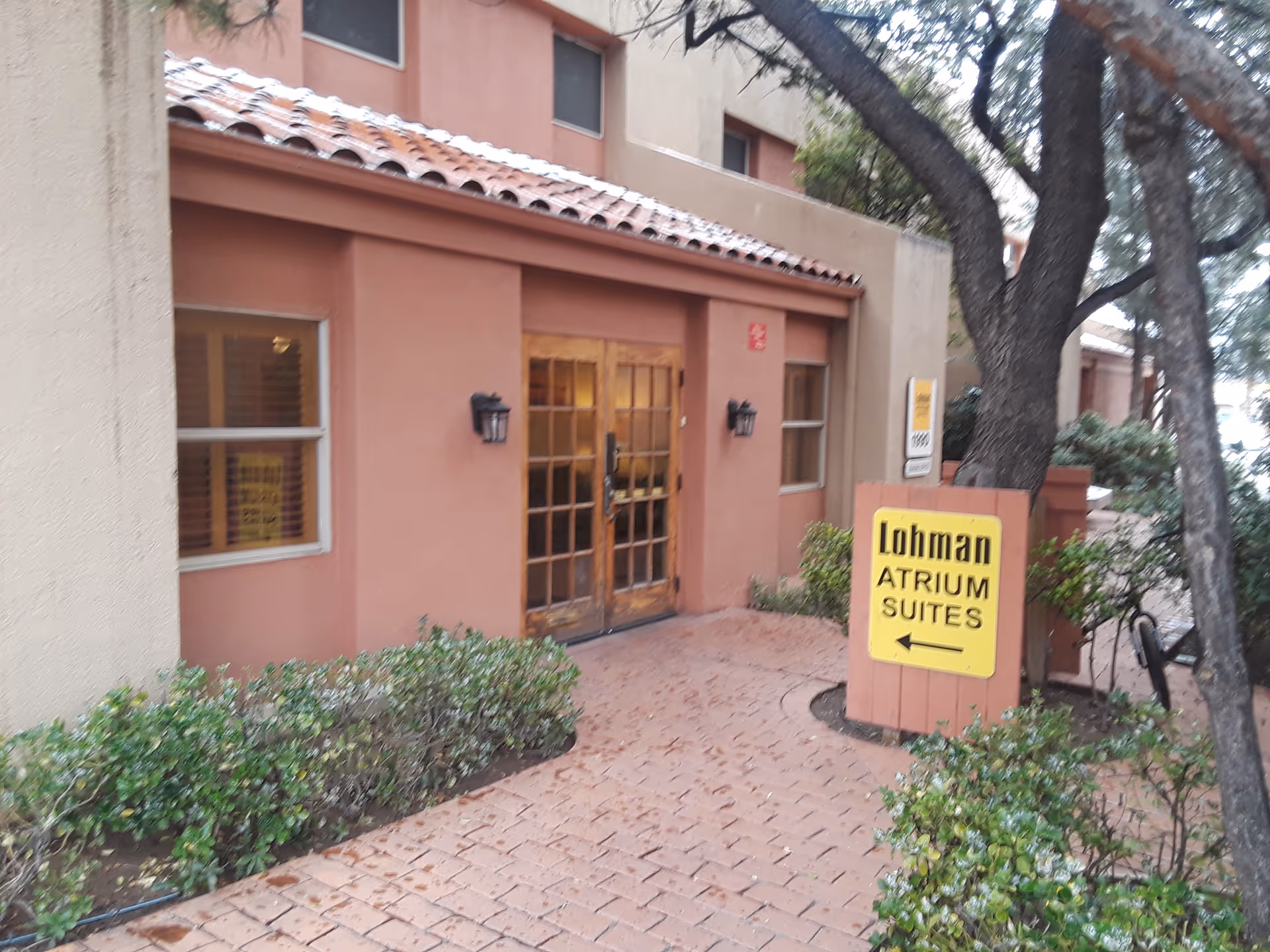 Exterior view of a beige building entrance with wooden doors and a yellow sign.