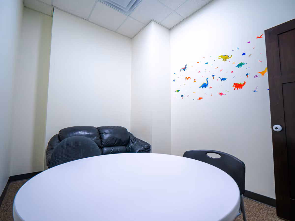 Small therapy room with round table, black chairs, a black leather sofa, and colorful dinosaur decals.