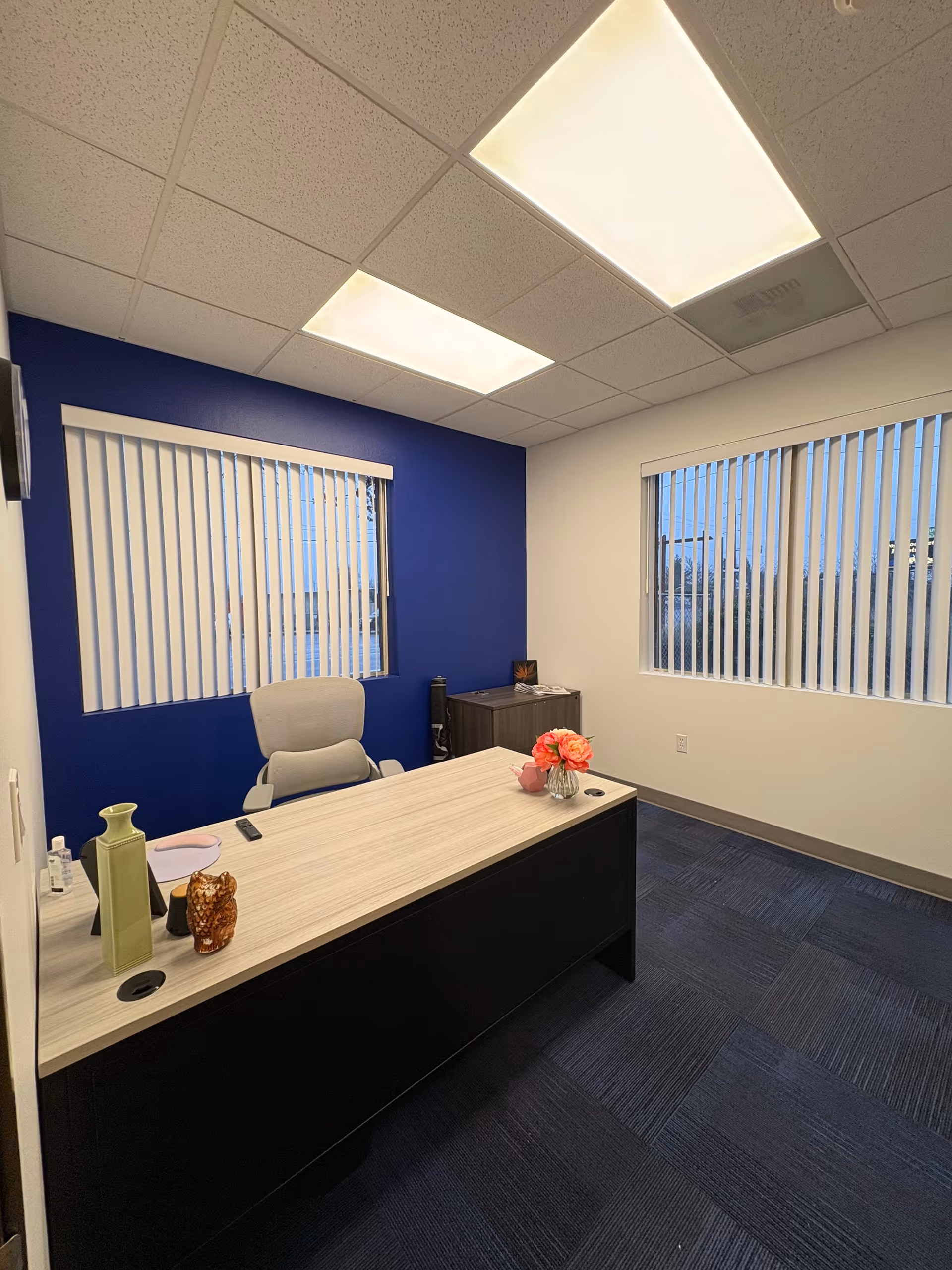 Office desk in a small room with blue accent wall, blinds, and overhead lights.