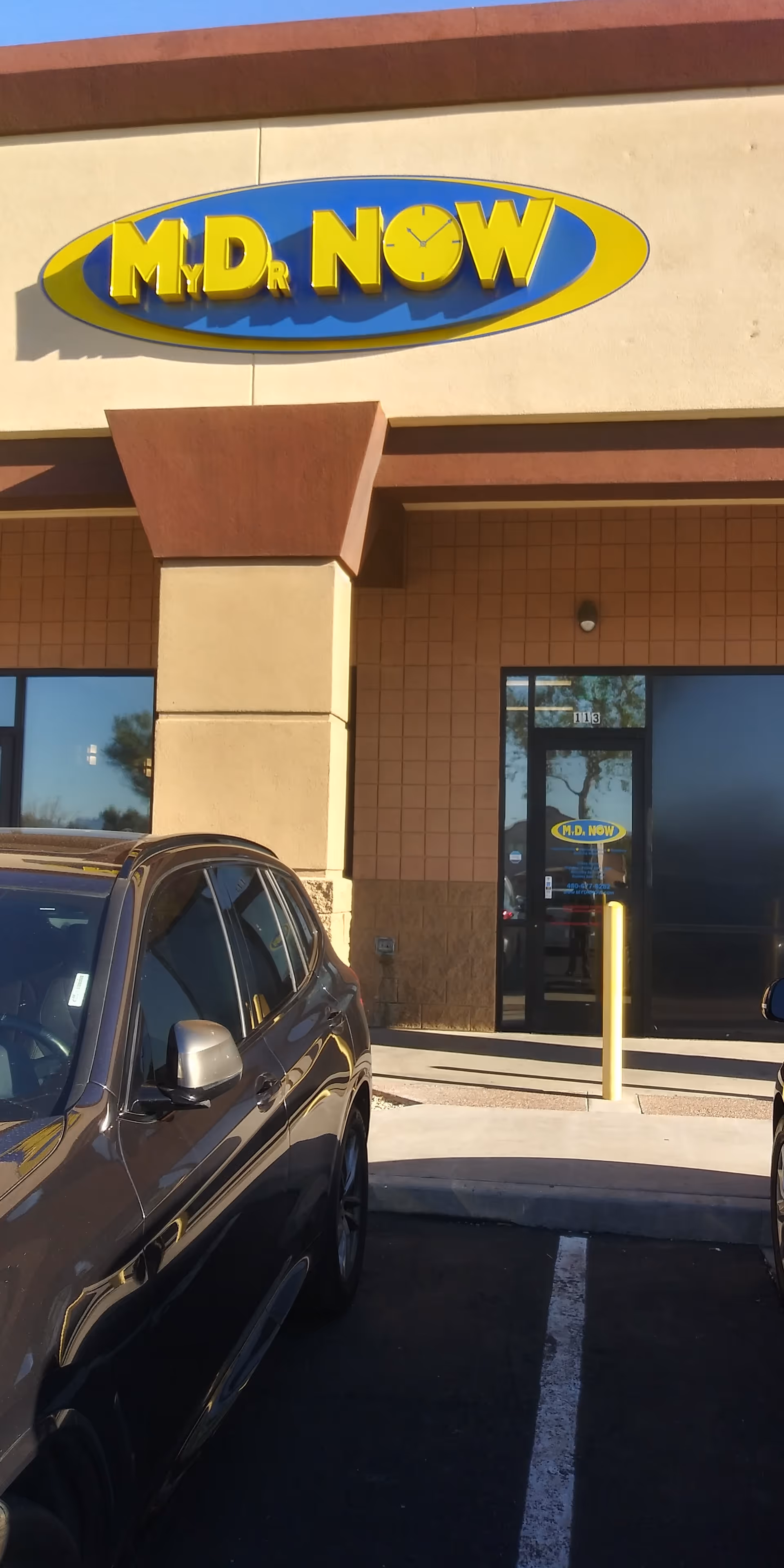 Front of a medical clinic with MD NOW sign, glass doors, and parked cars.