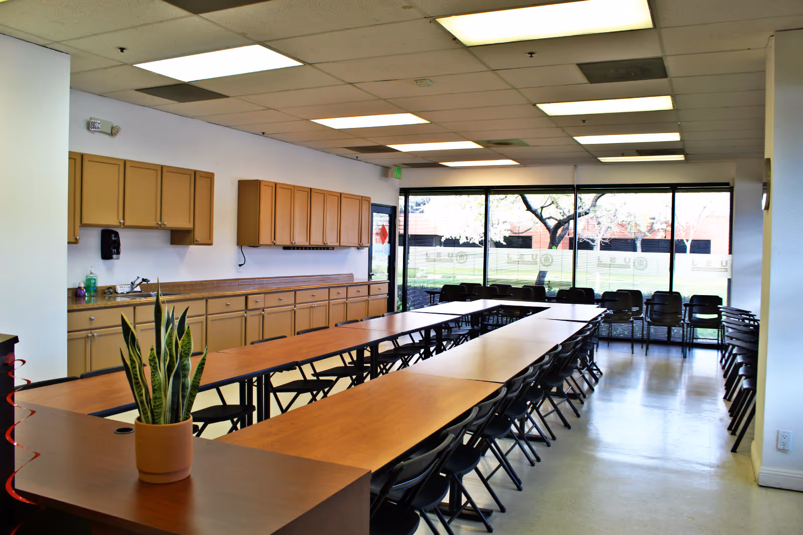 Large dining room with long tables, black chairs, wood cabinets, and large windows.