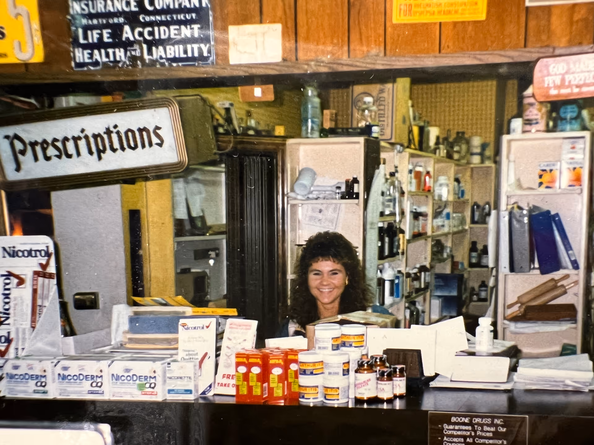 Smiling pharmacist behind a cluttered prescription counter with medicine bottles.