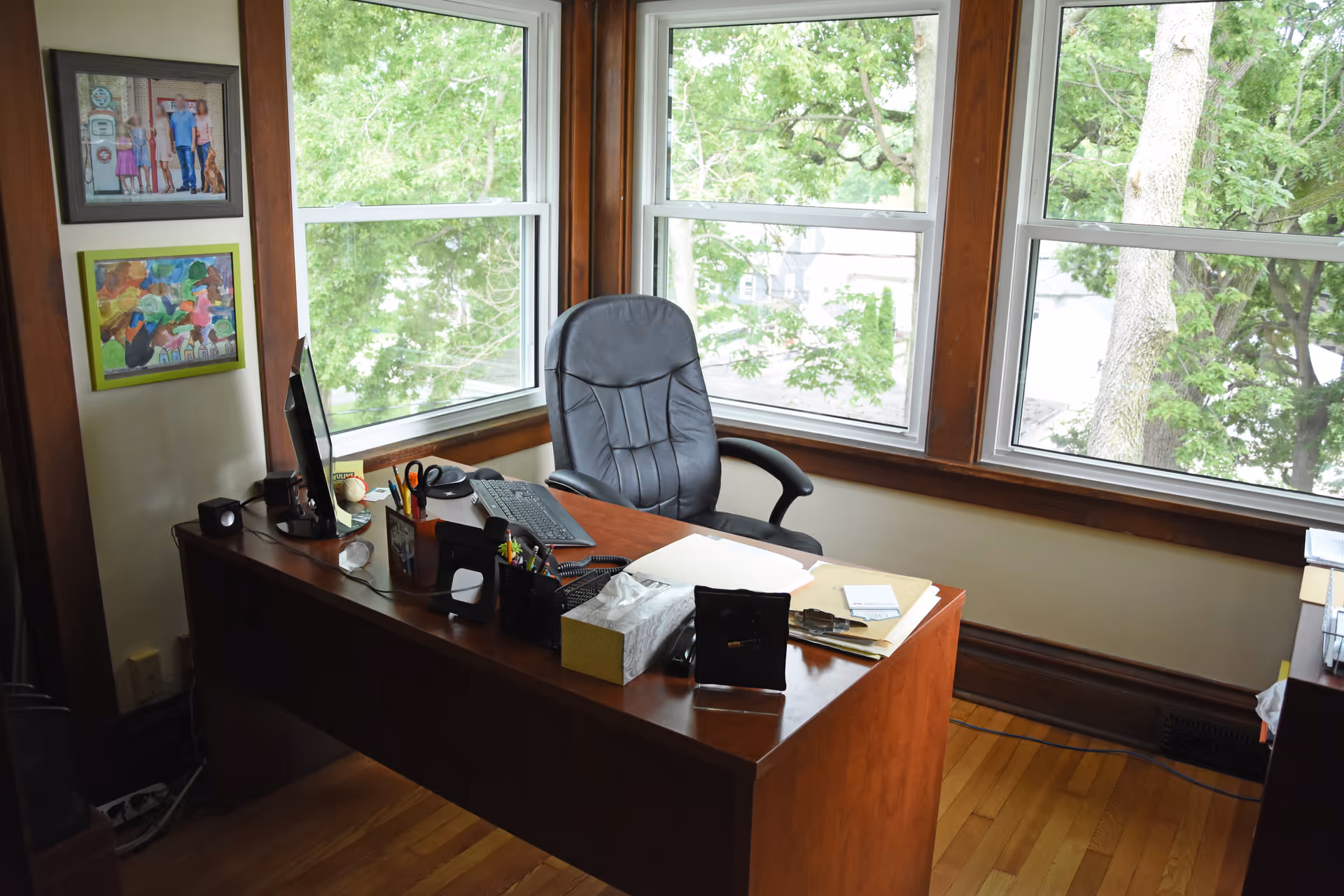 An organized wooden desk with computer, papers, and a high-back chair by large windows.