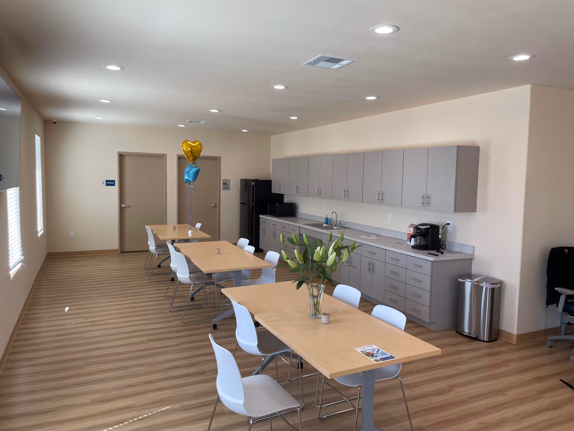 Bright communal dining area with tables, white chairs, and a kitchenette along the back wall.