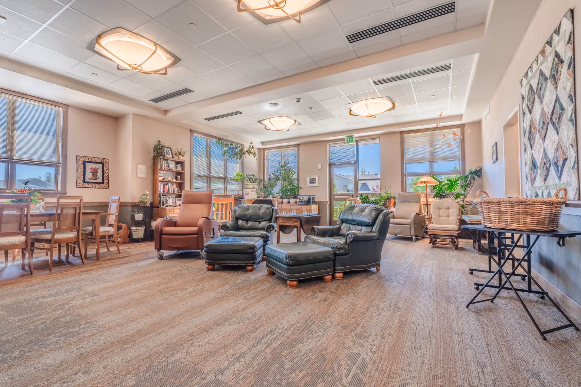 A spacious senior living lounge with recliners, chairs, plants, bookshelves, and large windows.