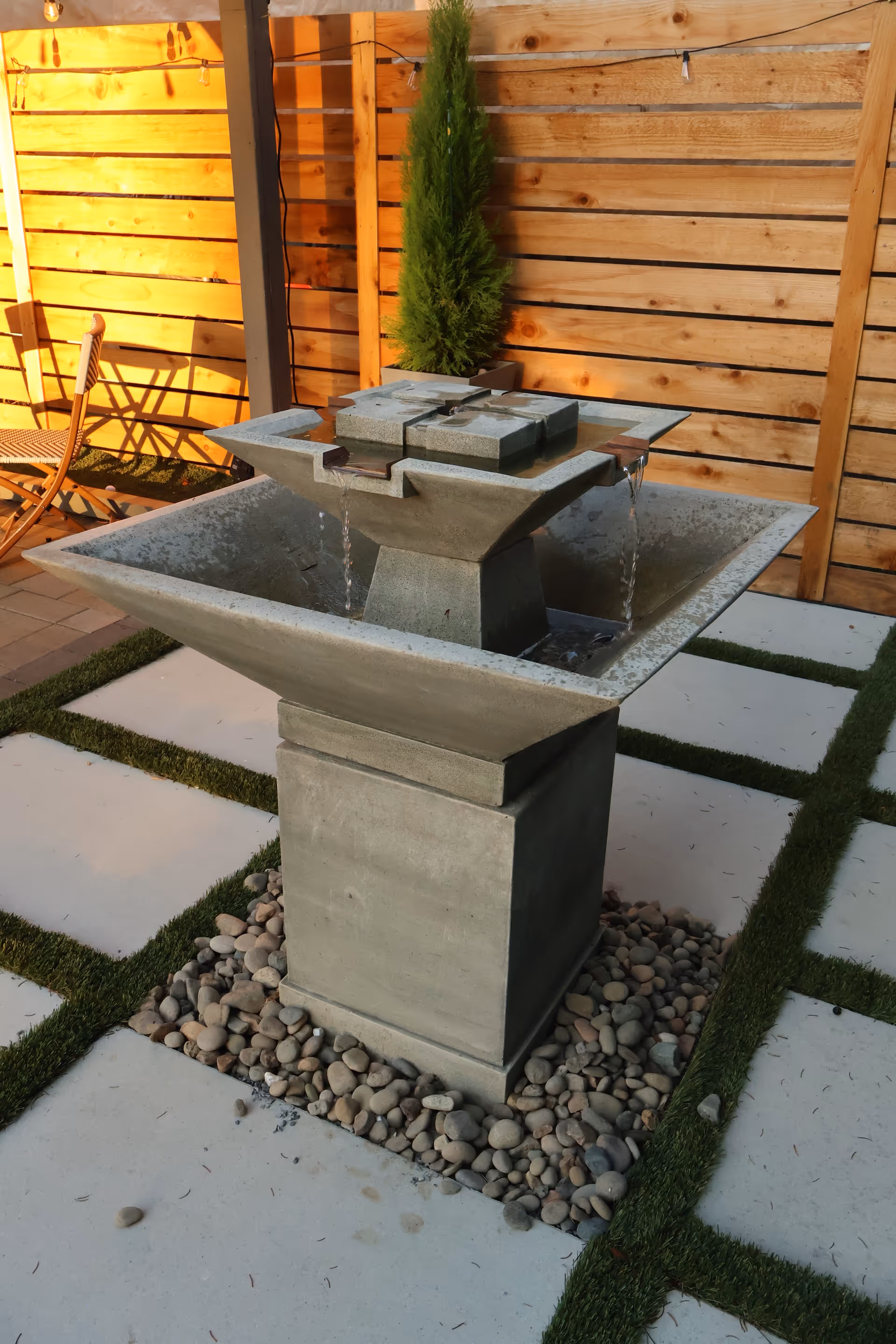 Outdoor courtyard with a modern stone water fountain, wooden fence, and pebble mulch.