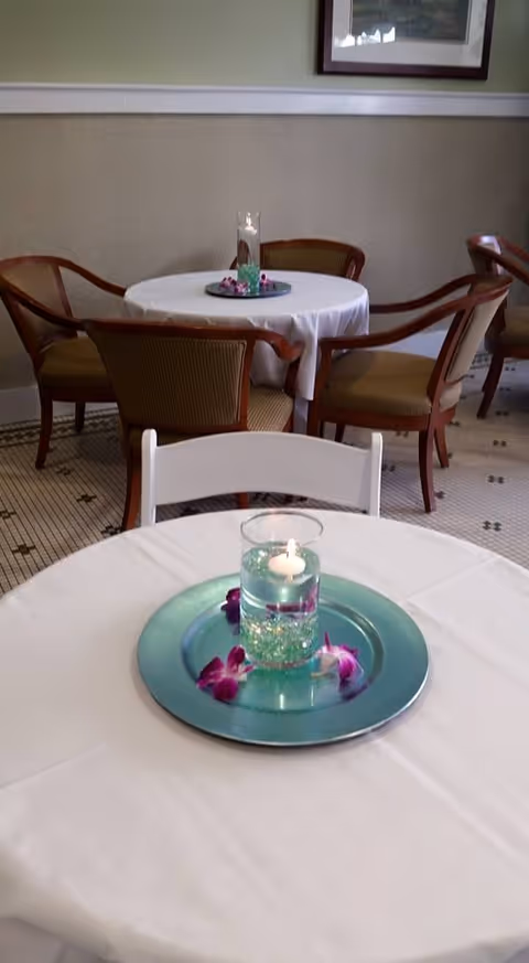 Round dining table with white cloth, teal centerpiece, and empty chairs in a dining room.