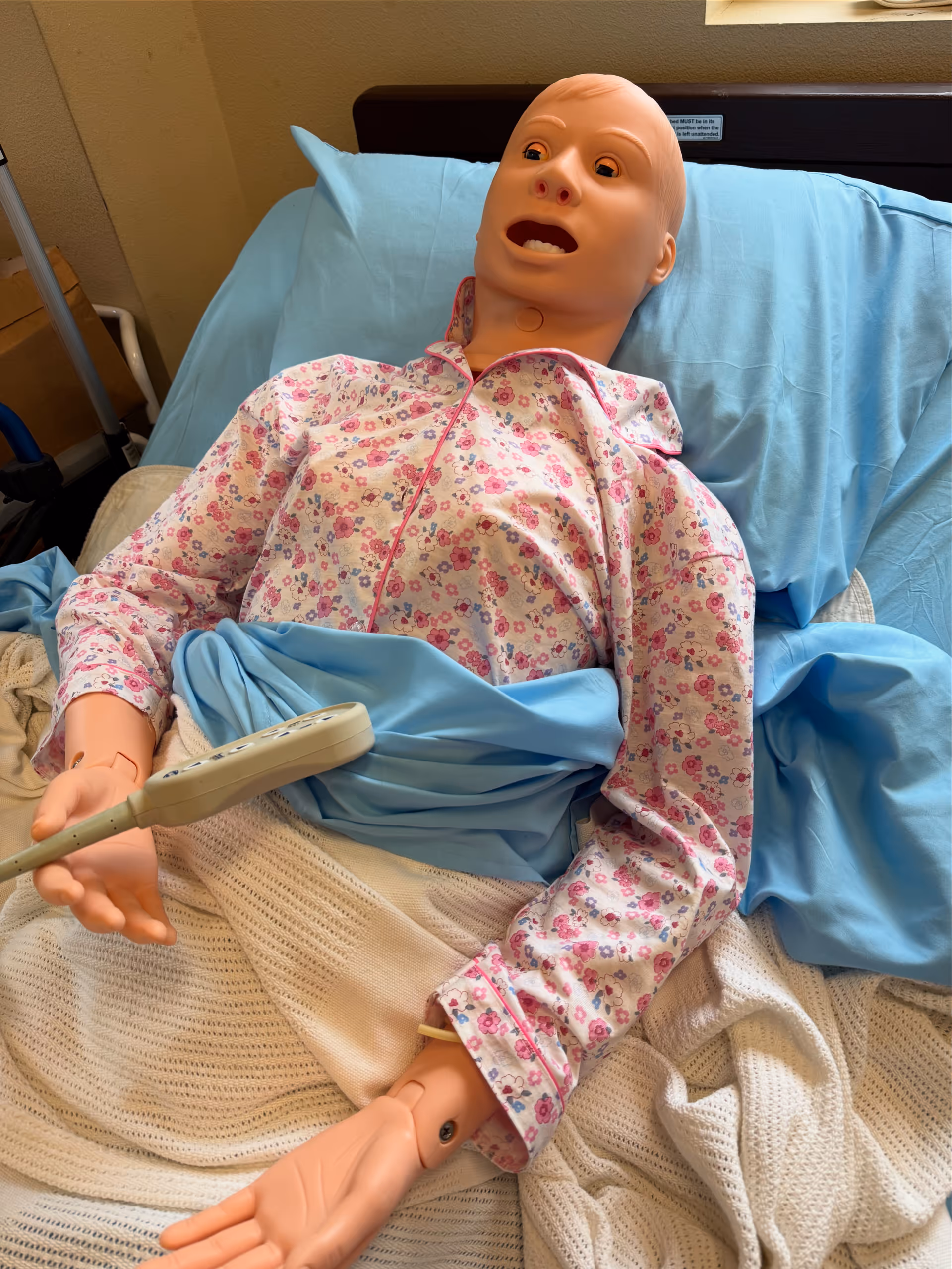A medical mannequin in a hospital bed wearing a floral gown.