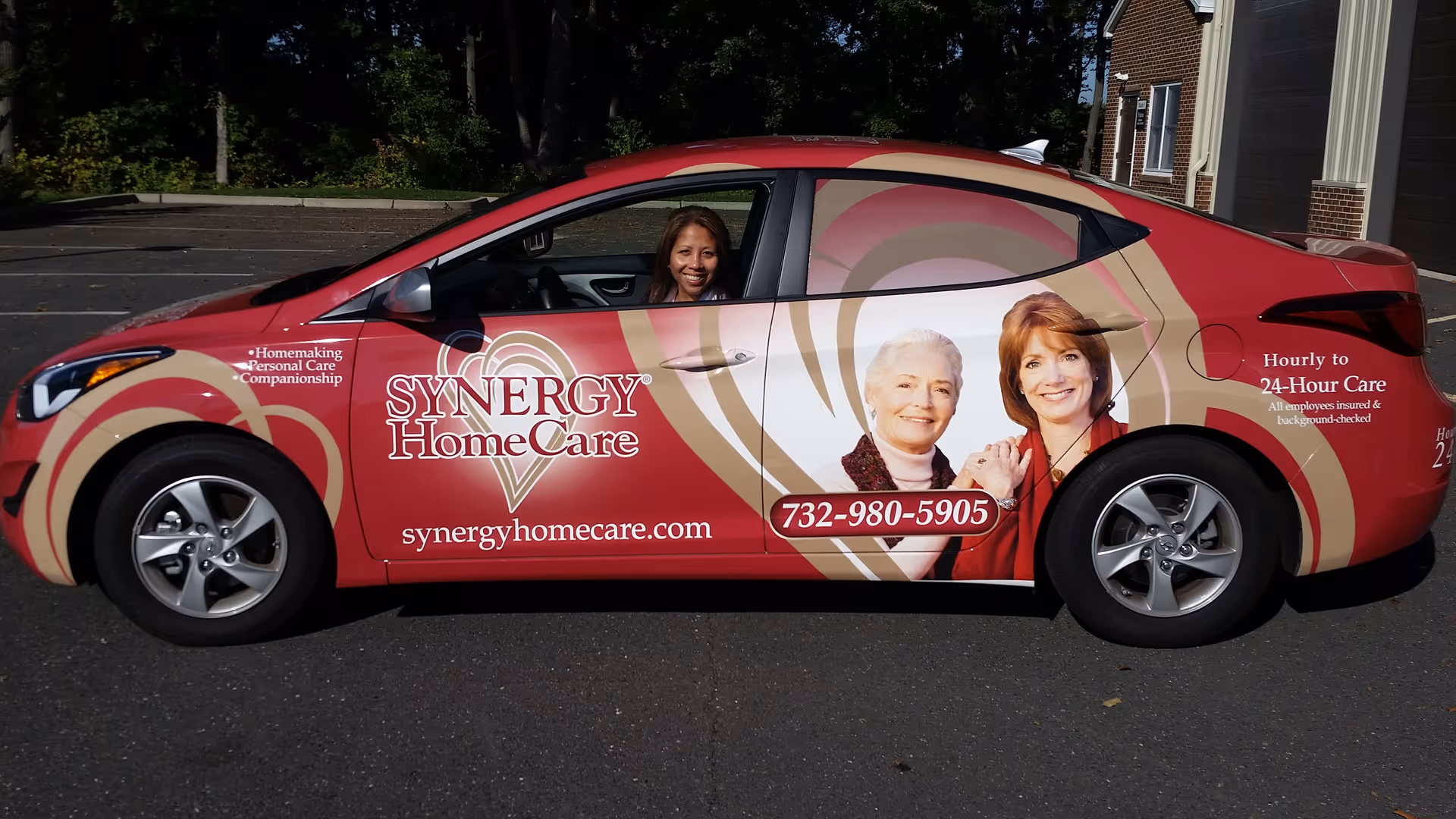 Red Synergy HomeCare car wrap parked outdoors with a smiling driver visible.