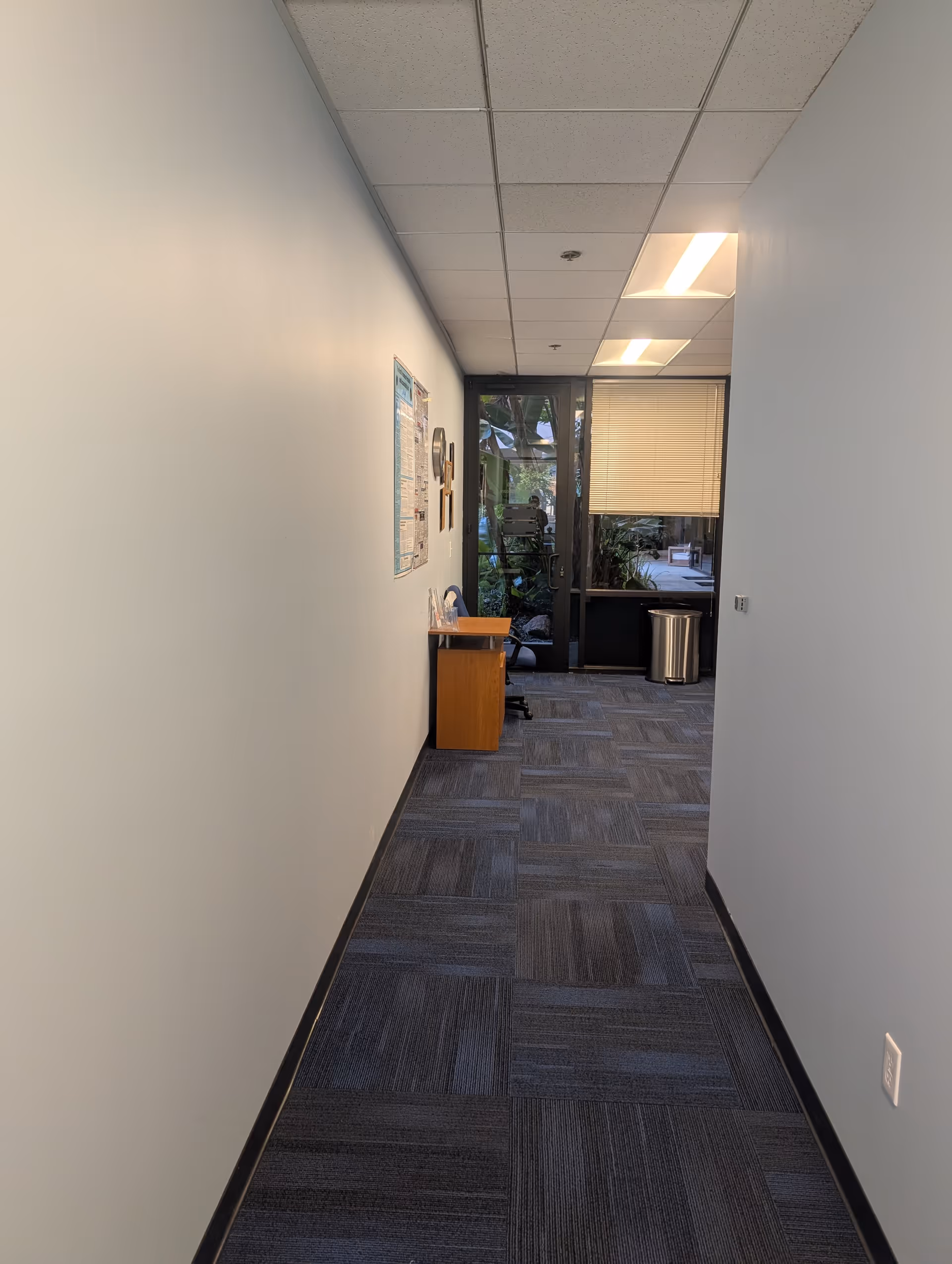 Narrow hallway with a bulletin board, small desk, and glass doors at the end.