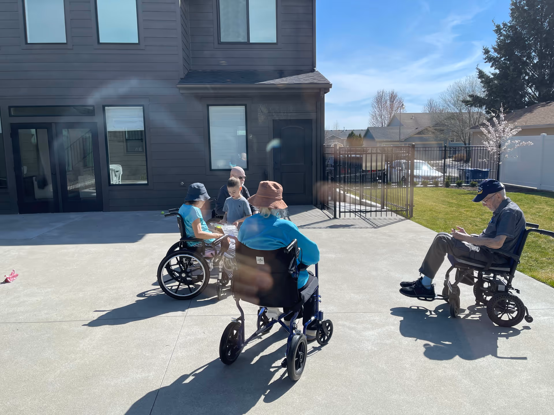 Residents in wheelchairs socialize outdoors beside a dark two-story building.