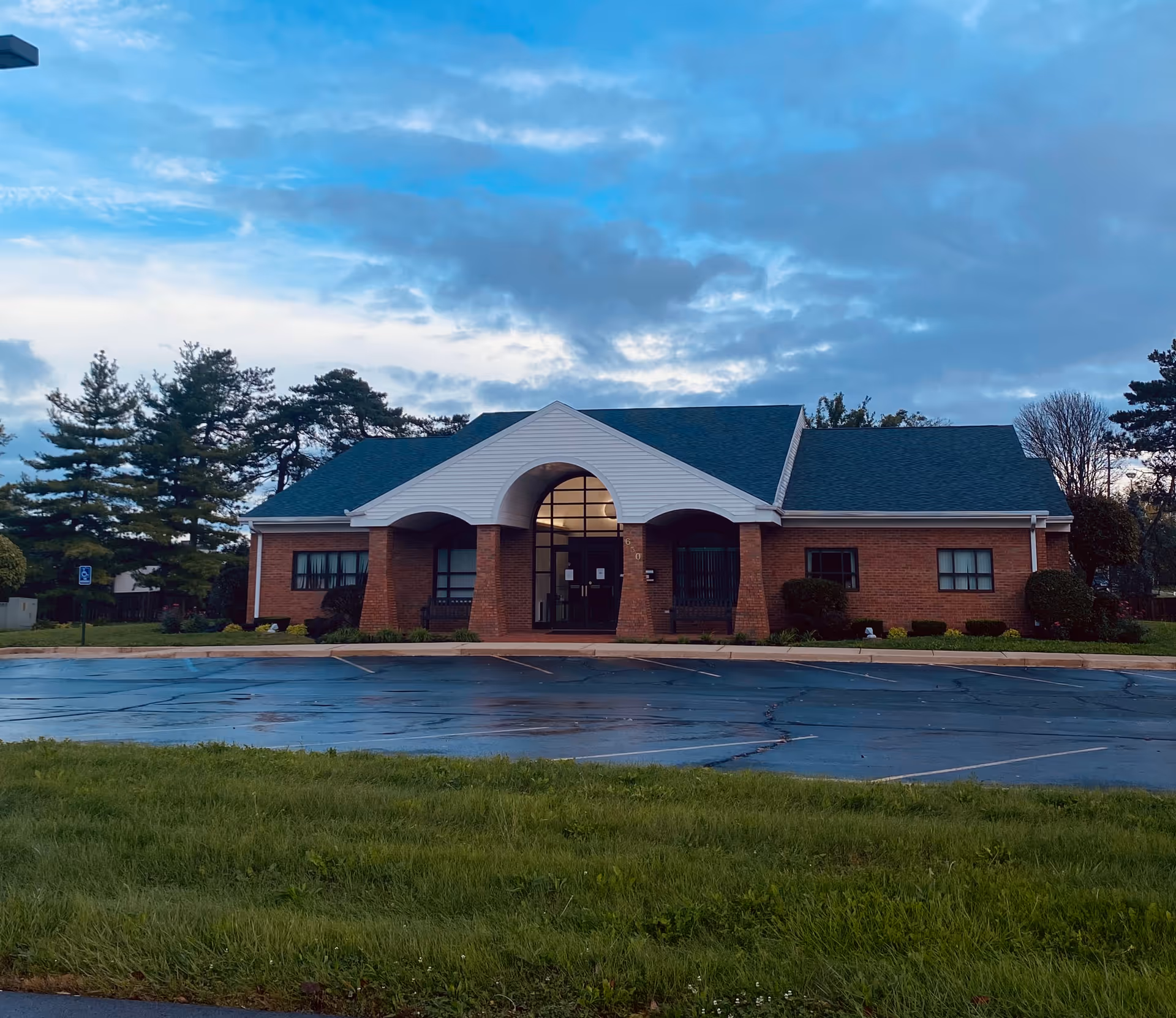 Brick senior living facility with arched entryway and surrounding greenery in a parking lot.