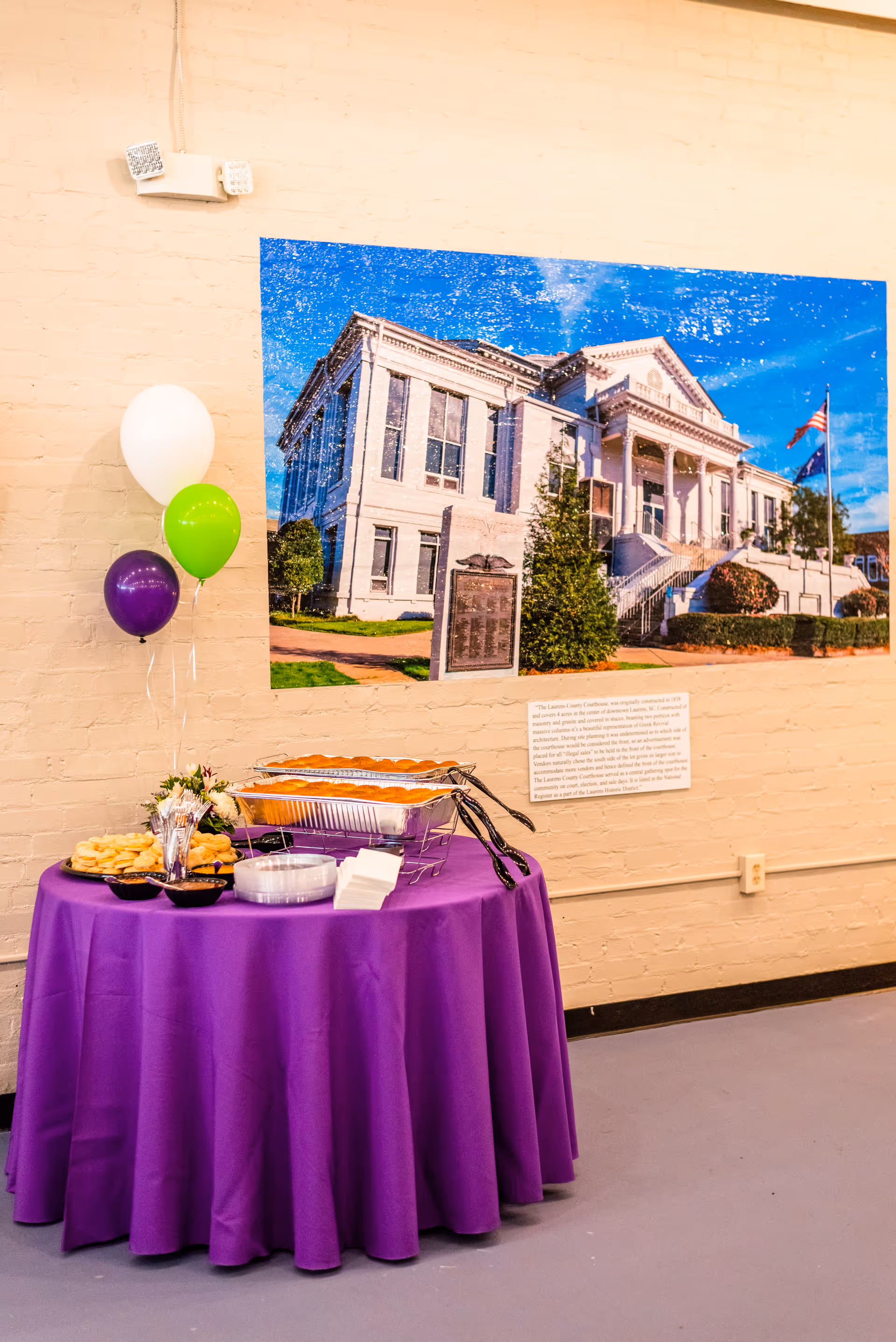 A purple-draped snack table with balloons sits indoors beside a large building photo.