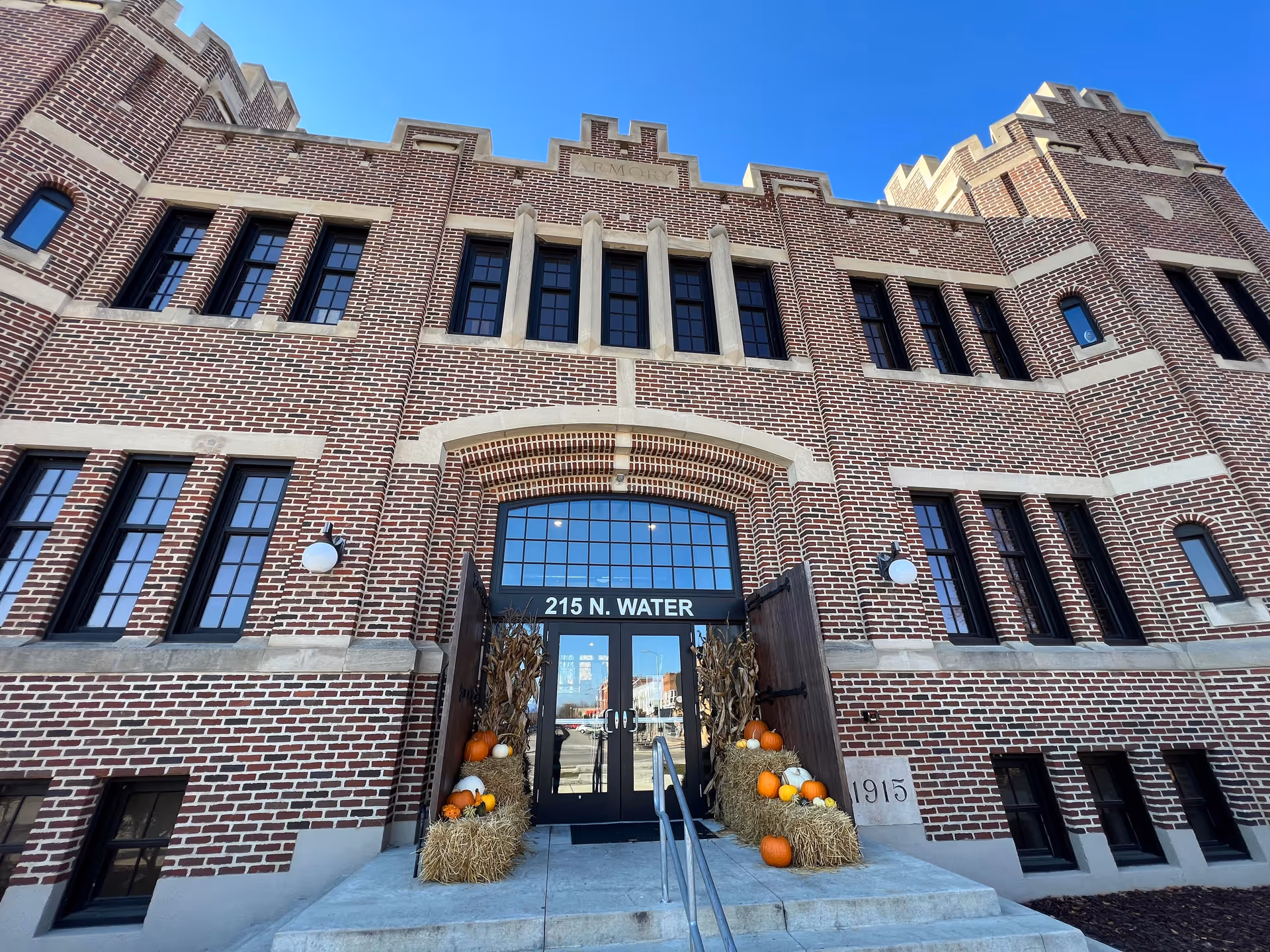 Brick senior home entrance with arched doors, pumpkins, hay bales, and a clear blue sky.