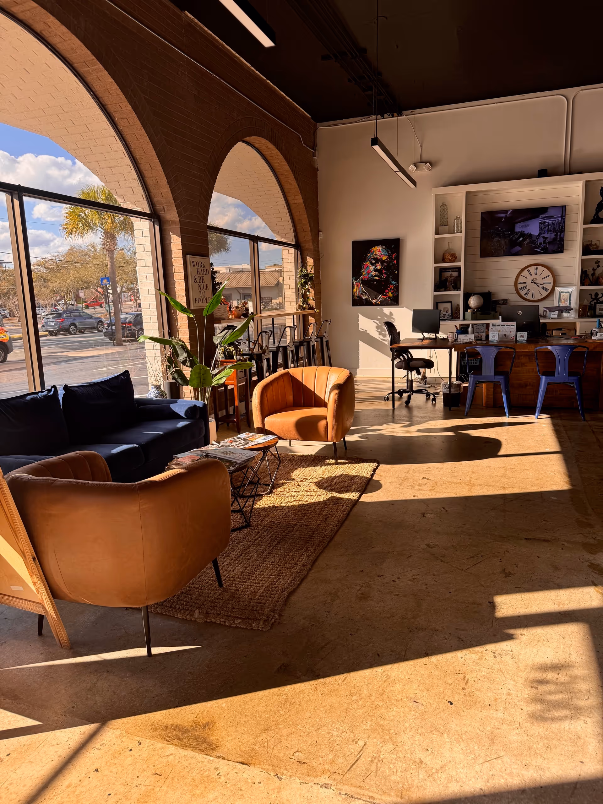 Sunlit interior lounge with tan chairs, rug, plants, and a desk near arches.