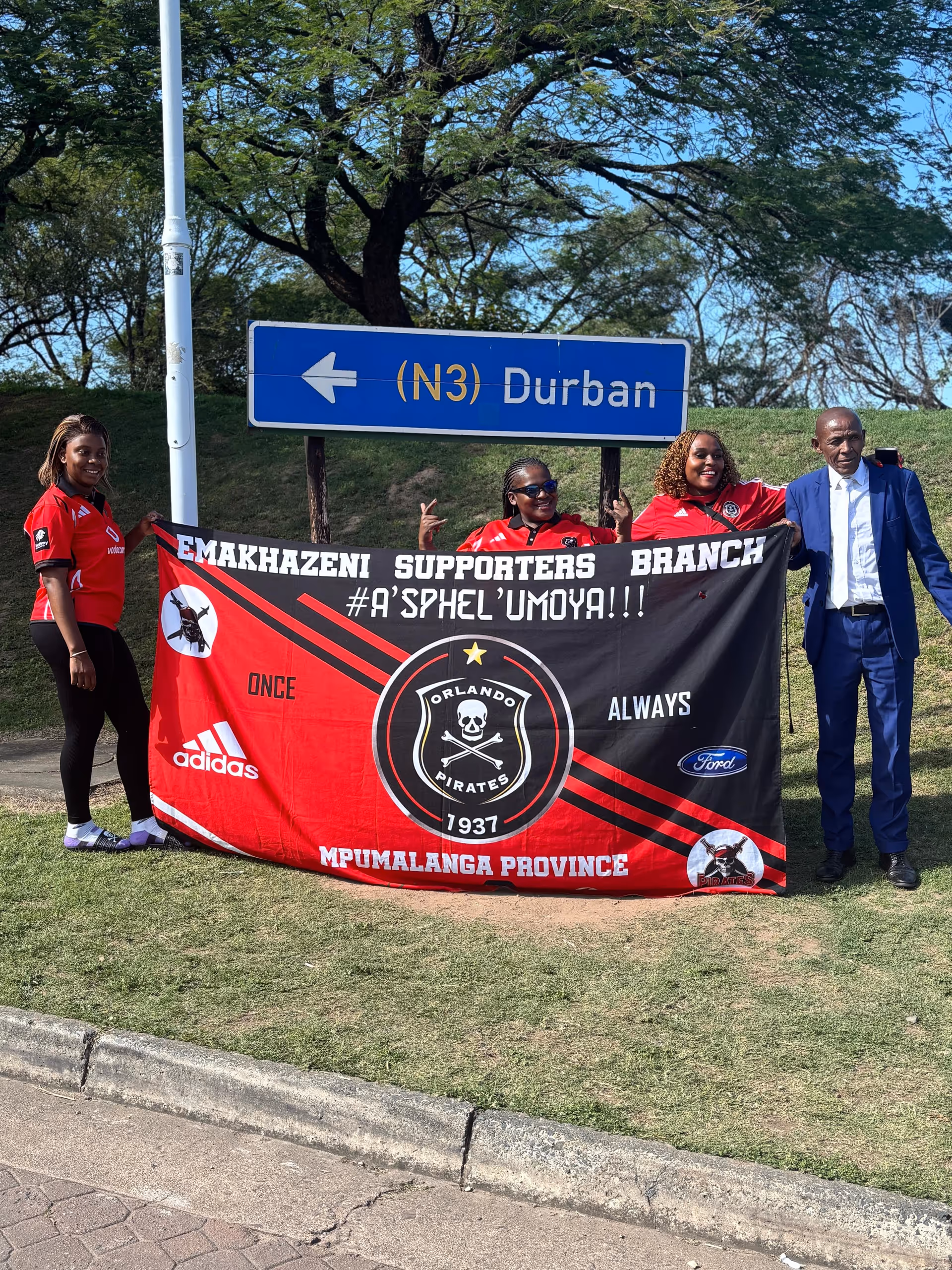 Four people hold a red and black banner beside a Durban road sign outdoors.