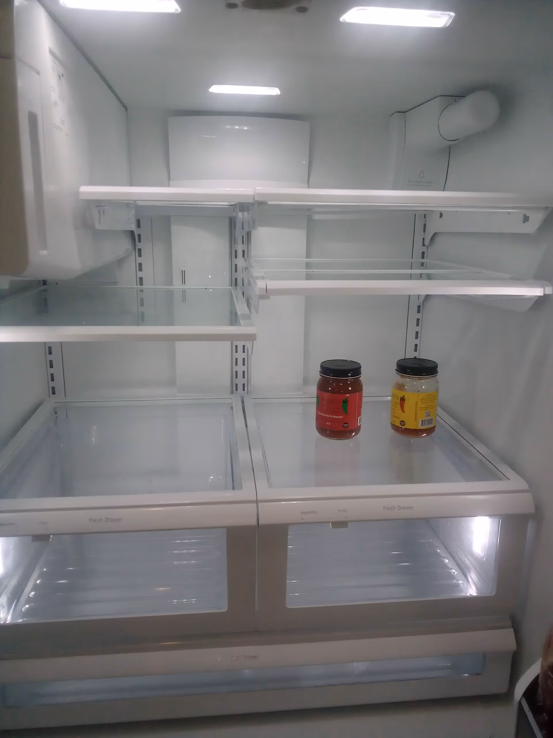 Open empty refrigerator in a facility kitchen with two jars on shelf.