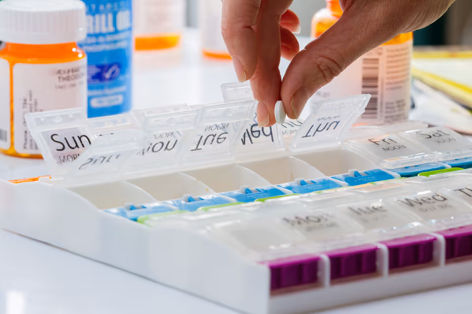 Close-up of a hand placing a pill into a weekly pill organizer on a table.