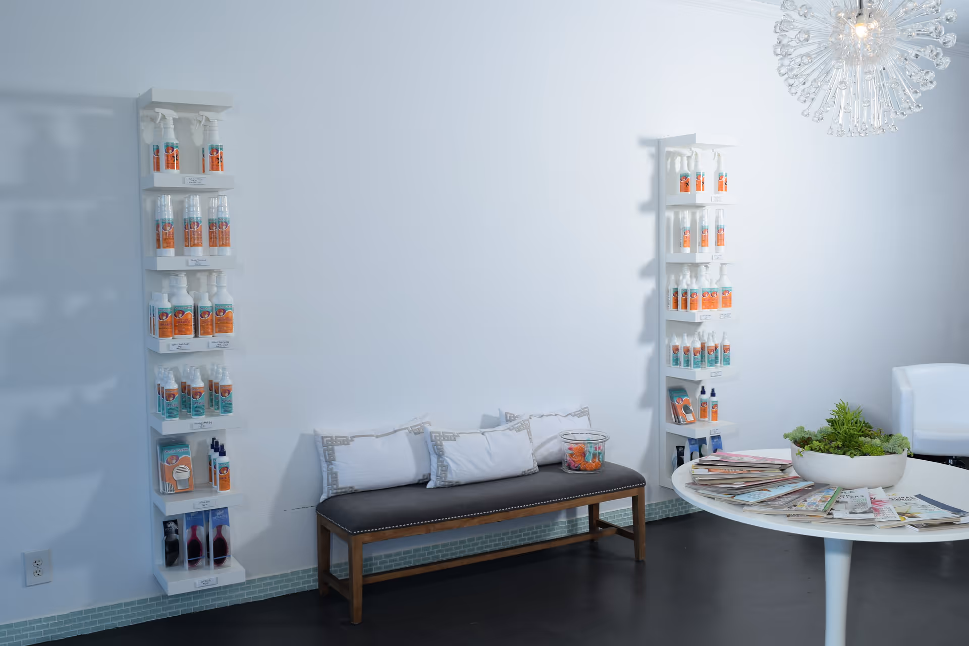 Minimalist lobby with white shelves of bottles, a cushioned bench, round table, magazines and a chandelier.