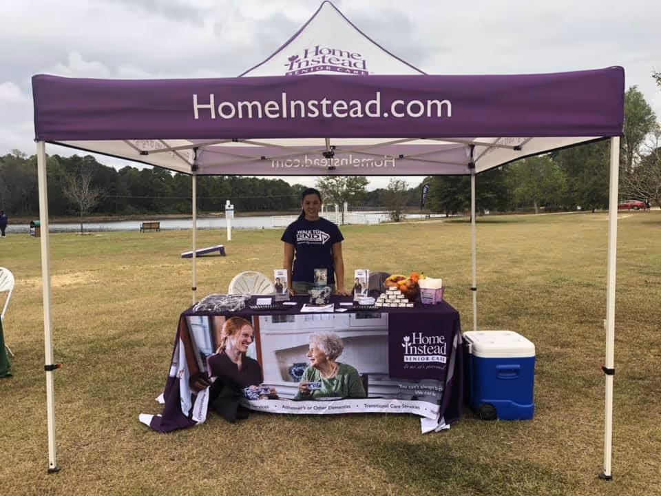 Staff member standing behind a purple Home Instead tent at an outdoor park by a lake.