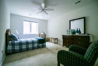 A furnished bedroom with a bed, dresser, chair, and window in a quiet, neutral room.