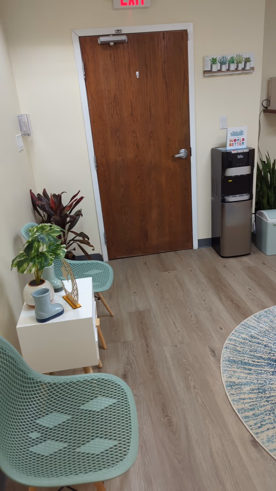Interior hallway with a wooden door, plants, and a water cooler near a round rug.