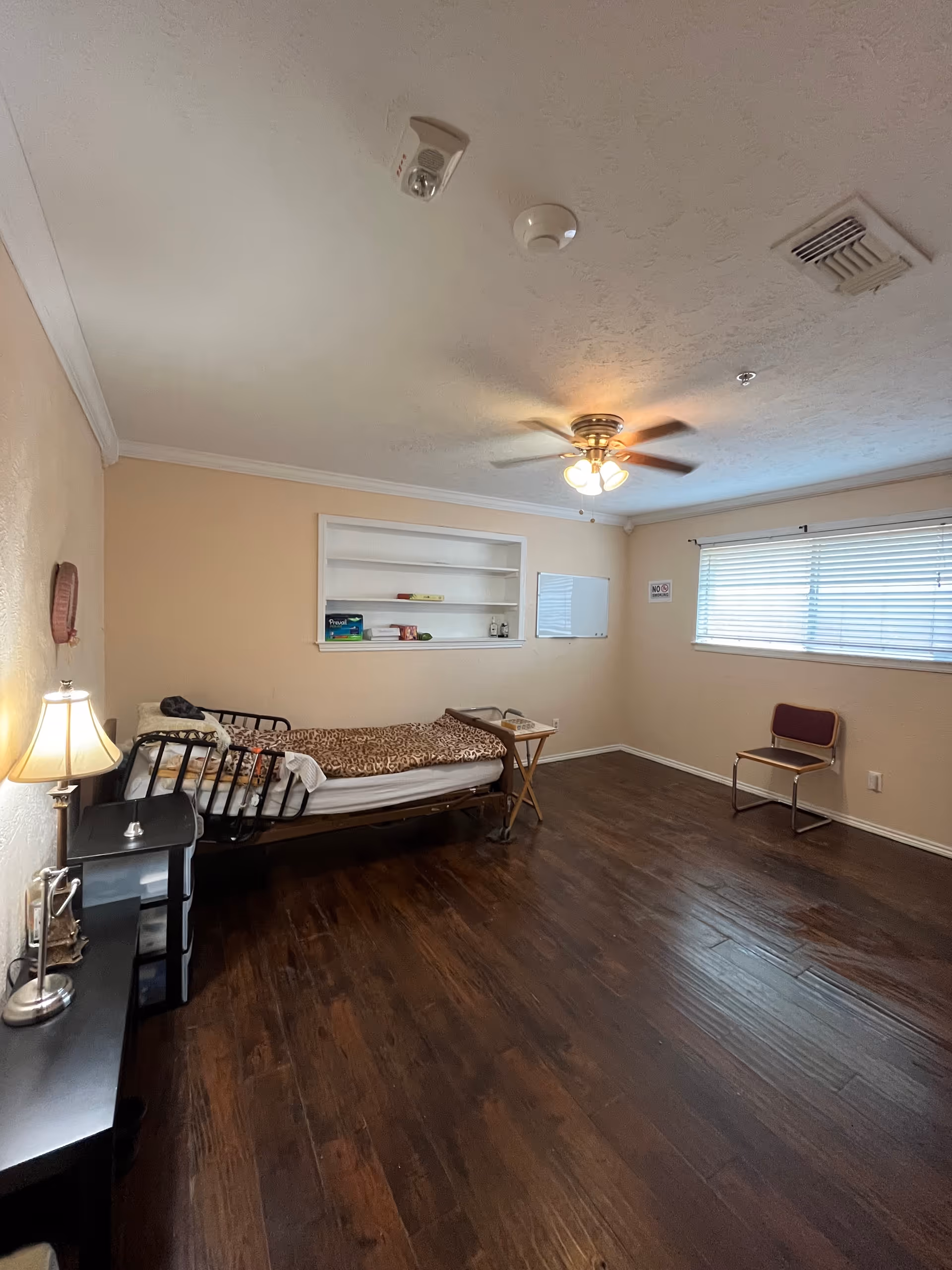 A sparsely furnished care-home bedroom with a bed, desk, shelf, chair, and window blinds.