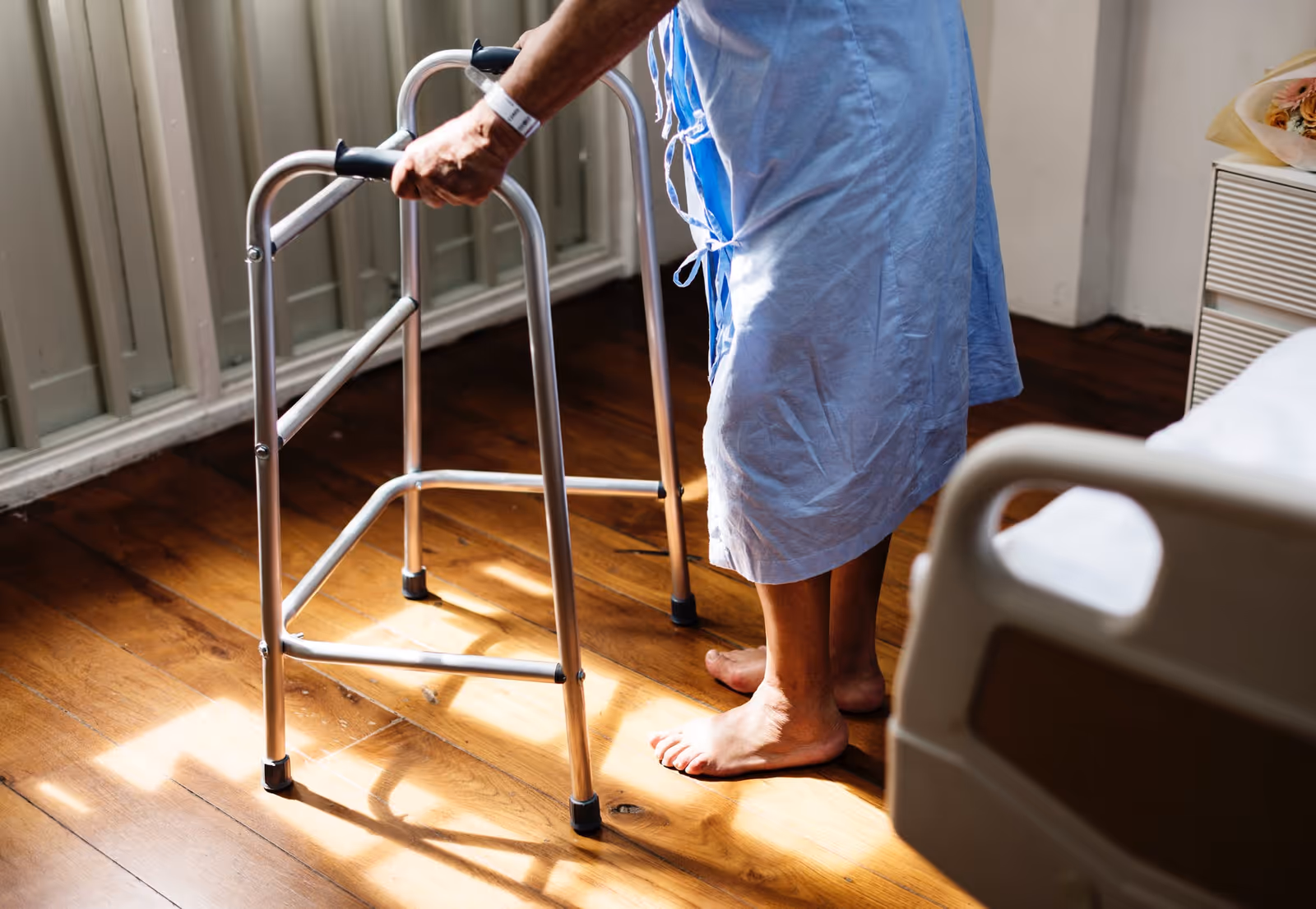 Senior resident uses a walker in a sunlit bedroom at a care facility.