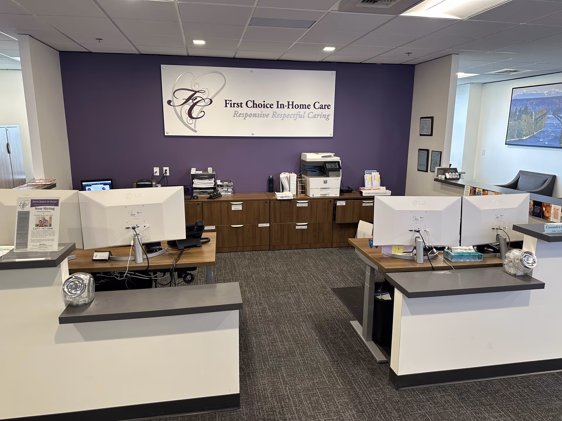 Reception area with dual desks, computers, and a purple wall sign reading First Choice In-Home Care.